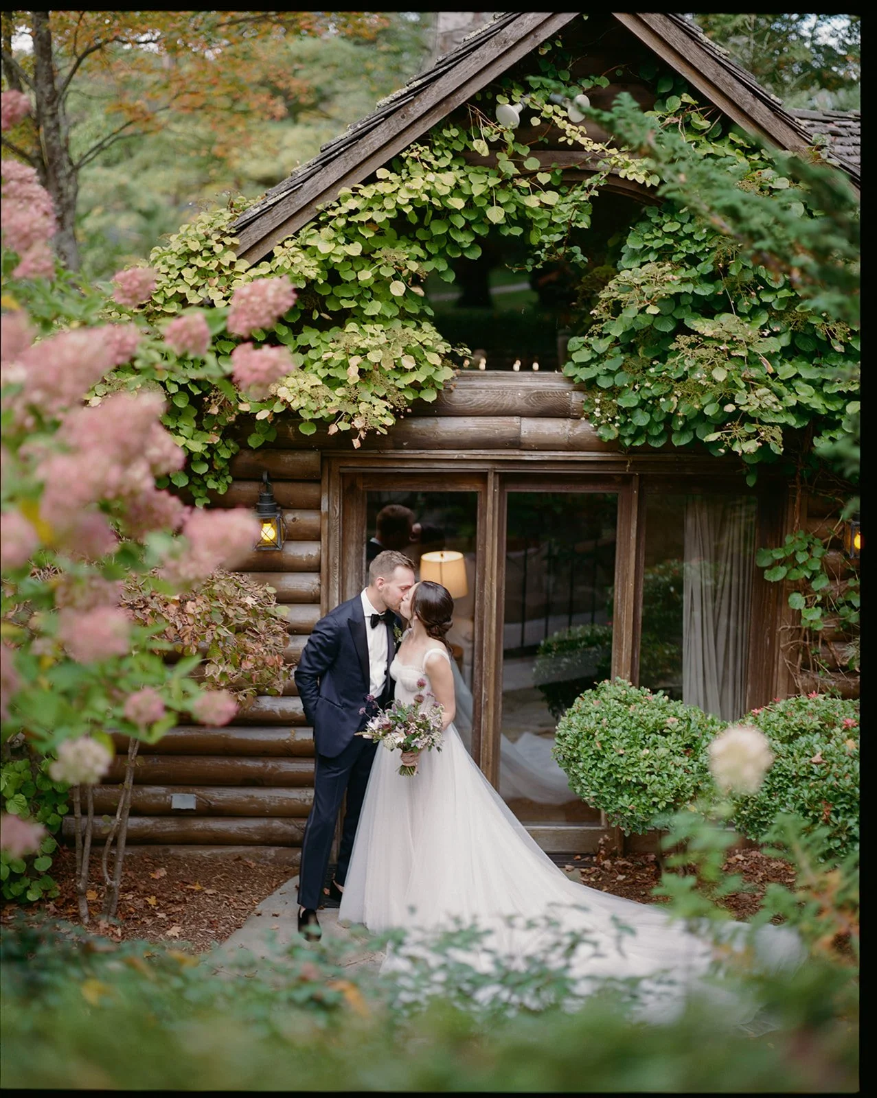 A bride and groom sharing a kiss outside a rustic wooden cabin, surrounded by lush green foliage and pink flowers.
