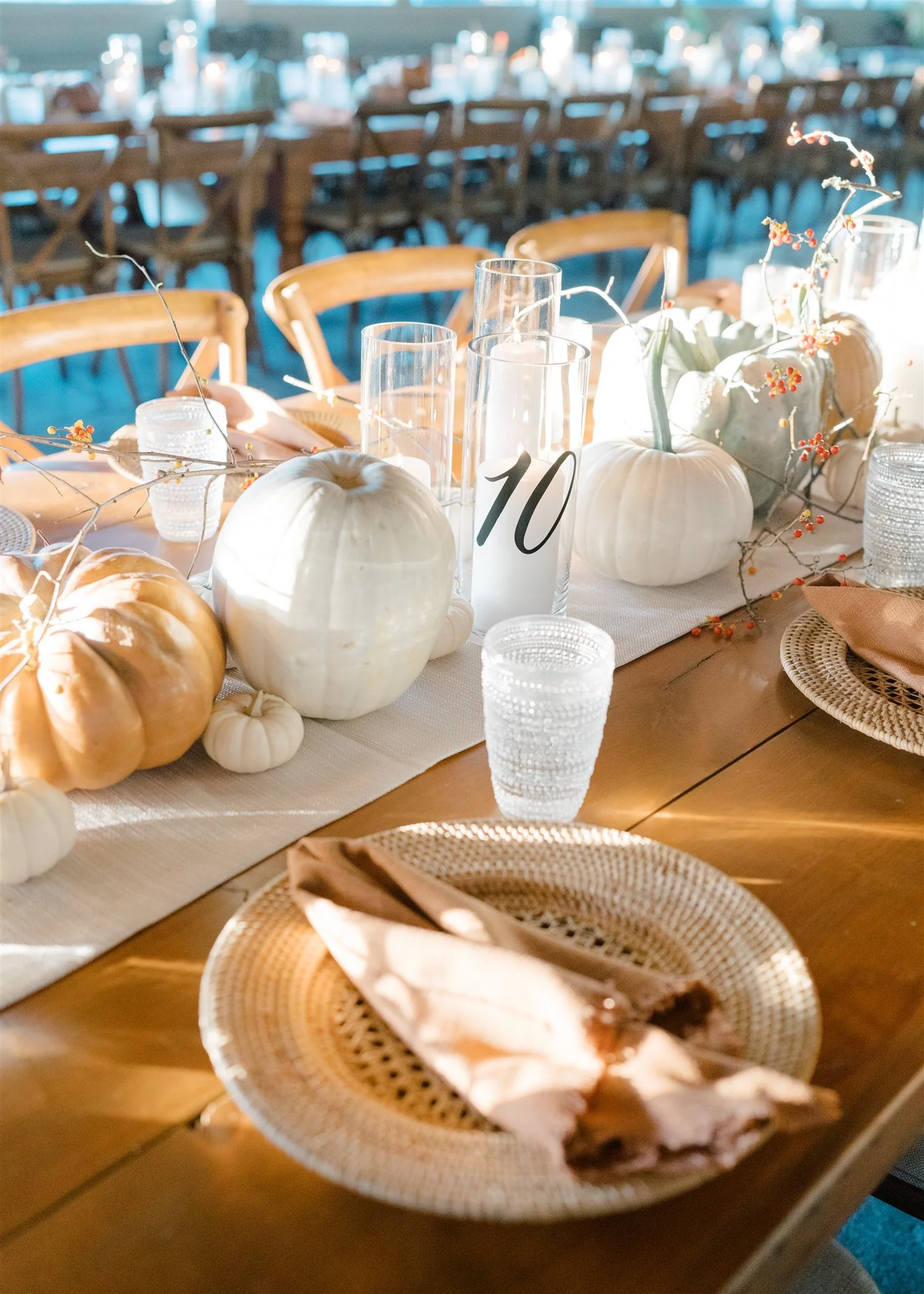 A decorated table with fall-themed pumpkins, branches with berries, glass candle holders, a table number '10', woven placemats, folded napkins, and glassware, set up for a celebration or event.