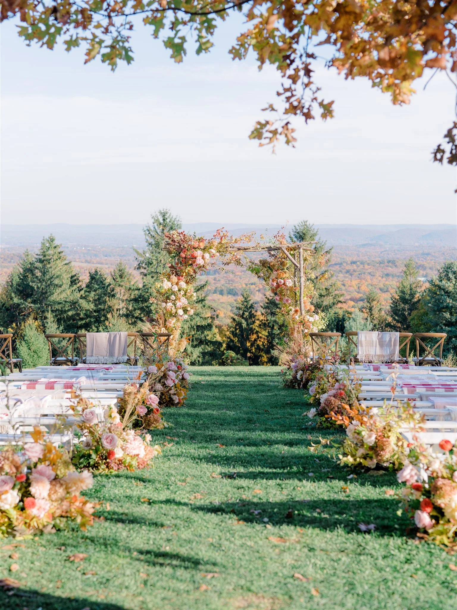 An outdoor wedding ceremony setup on a grassy hill with rows of white chairs and floral arrangements along the aisle leading to a wooden arch adorned with flowers, overlooking a scenic landscape of trees and mountains.