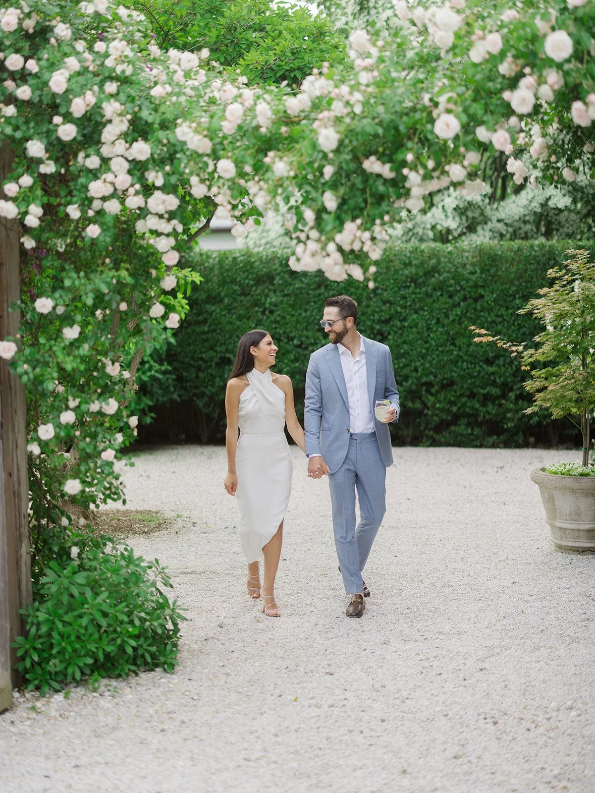 A couple dressed in formal attire, holding hands and walking outdoors on a gravel path, surrounded by lush green trees and blooming white flowers.