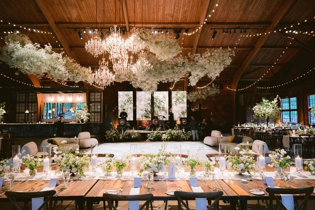 Elegant wedding reception with a long wooden table decorated with white flowers, candles, and tableware, set in a rustic barn with a wooden ceiling, string lights, hanging chandeliers, floral arrangements, a dance floor, and a band stage in the backg