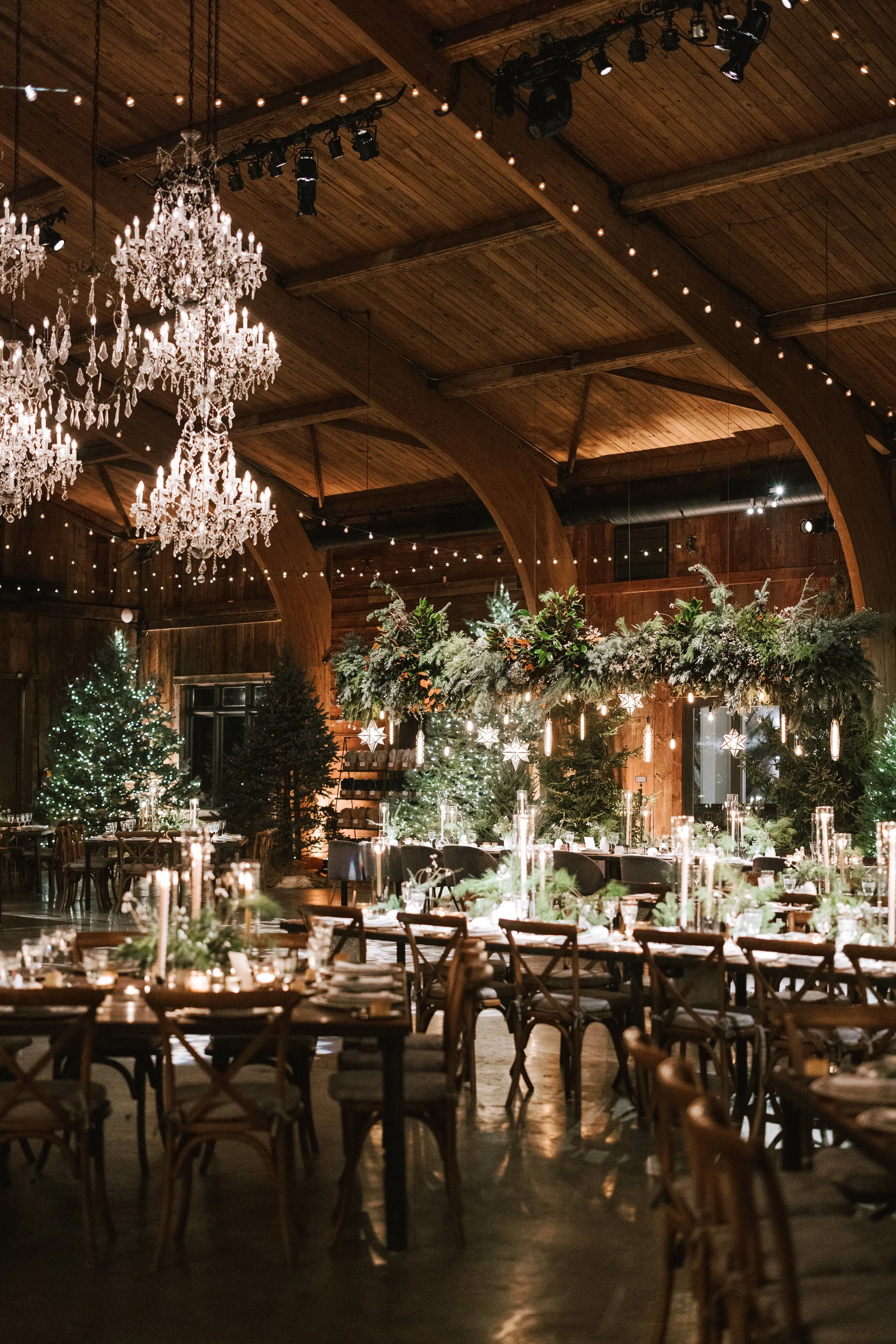 Festively decorated indoor venue with wooden ceiling and walls, adorned with chandeliers, string lights, and Christmas trees, set up for a holiday celebration with tables, chairs, and elegant table settings.