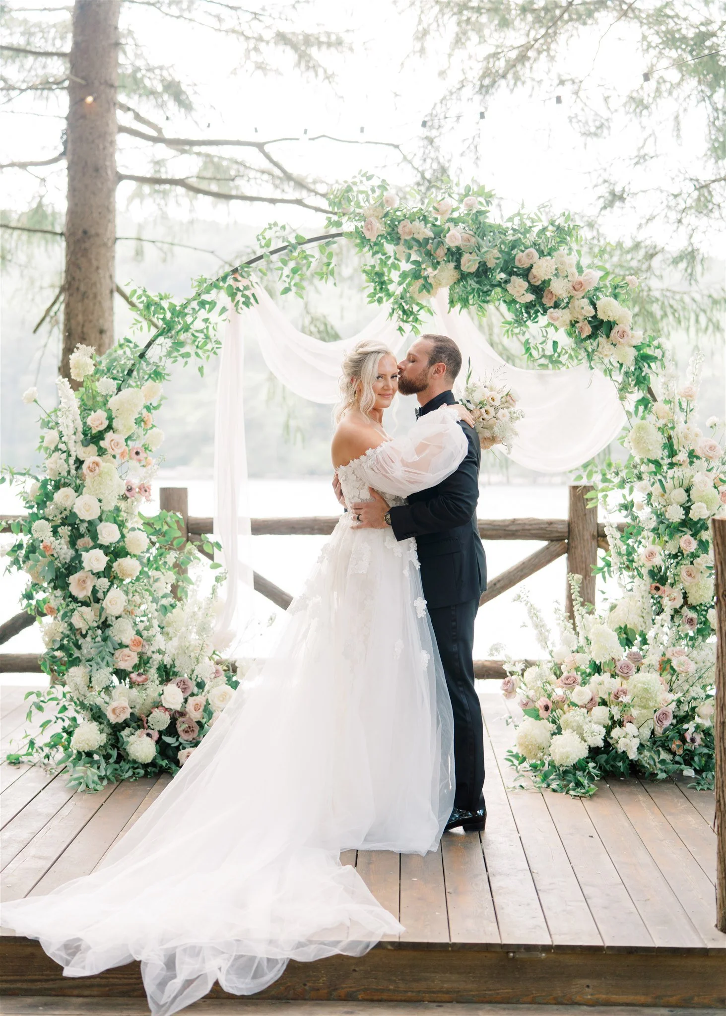 A bride and groom sharing a kiss on an outdoor wooden platform with a scenic lake background, surrounded by a floral archway of white and pink roses and greenery.