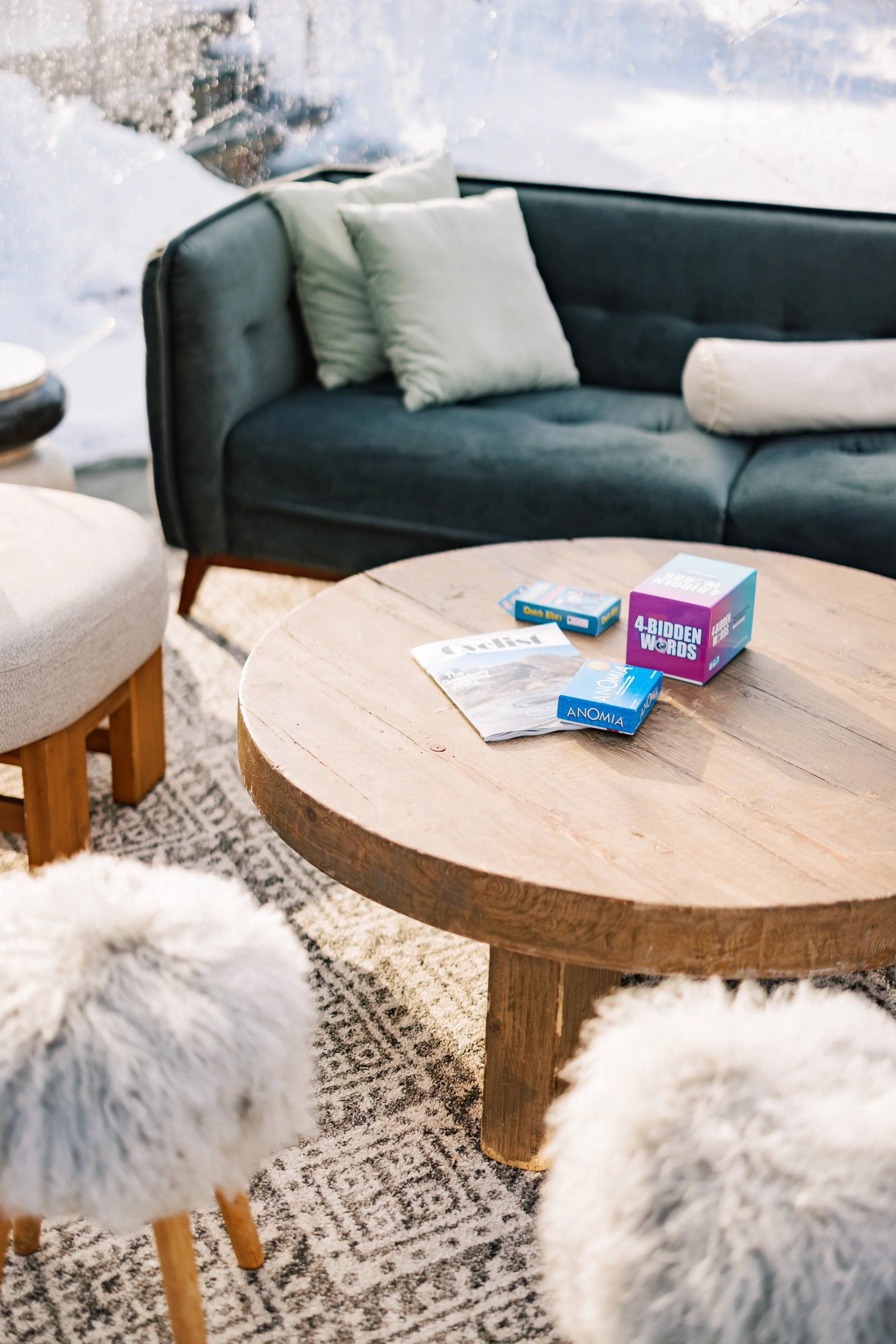 A round wooden coffee table with puzzle books and game boxes on it, surrounded by a dark blue sofa with pillows, a beige and fur-covered stool, and a patterned rug. Snow is visible outside through a window.