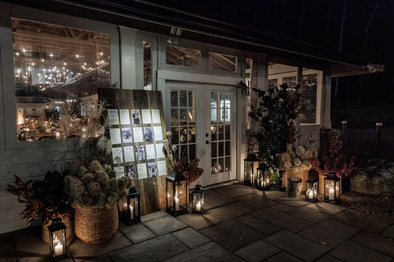 Nighttime exterior of a decorated venue entrance with lanterns, floral arrangements, and a seating chart displayed on a wooden board.