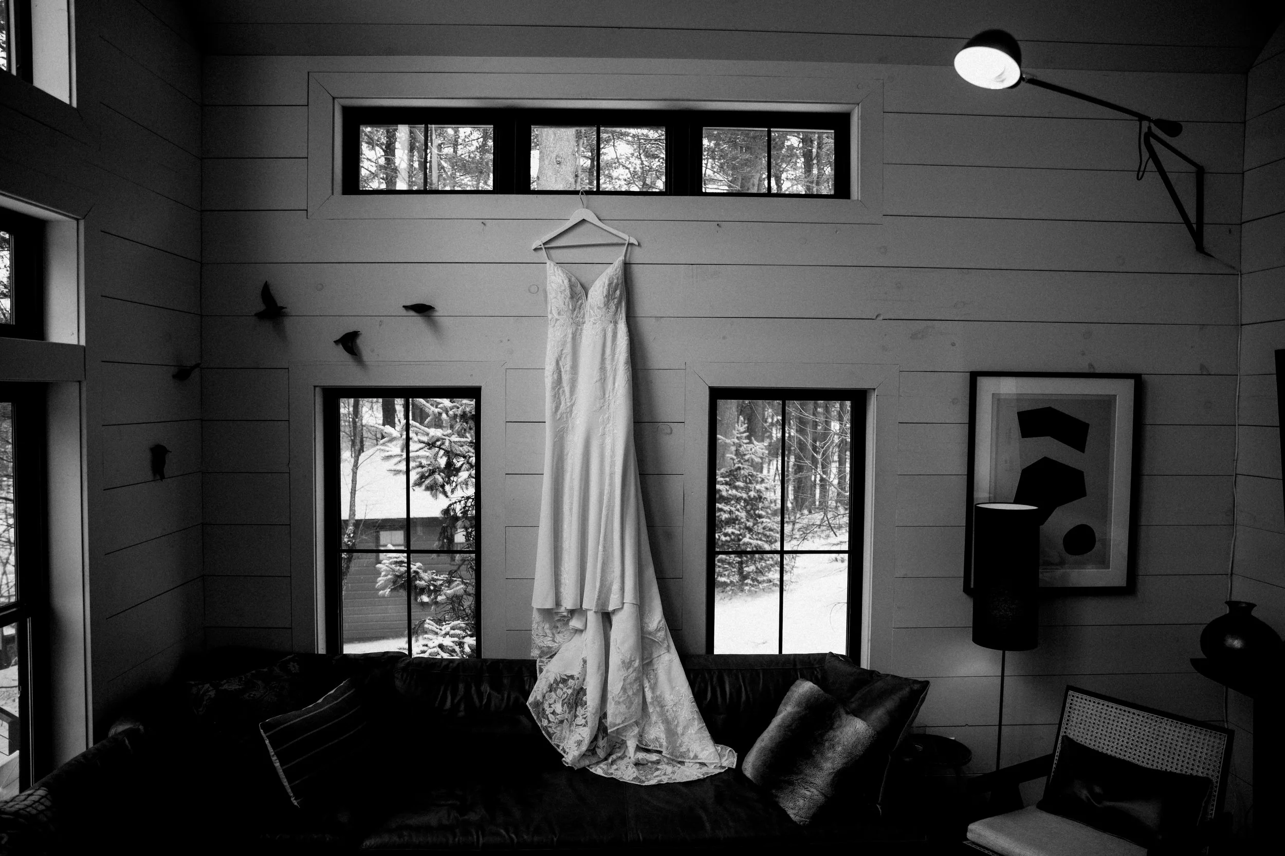 A white wedding dress hanging on a wooden wall with two large windows behind it showing snow outside. The room contains a sofa with pillows, a framed abstract art piece, a floor lamp, and a chair.