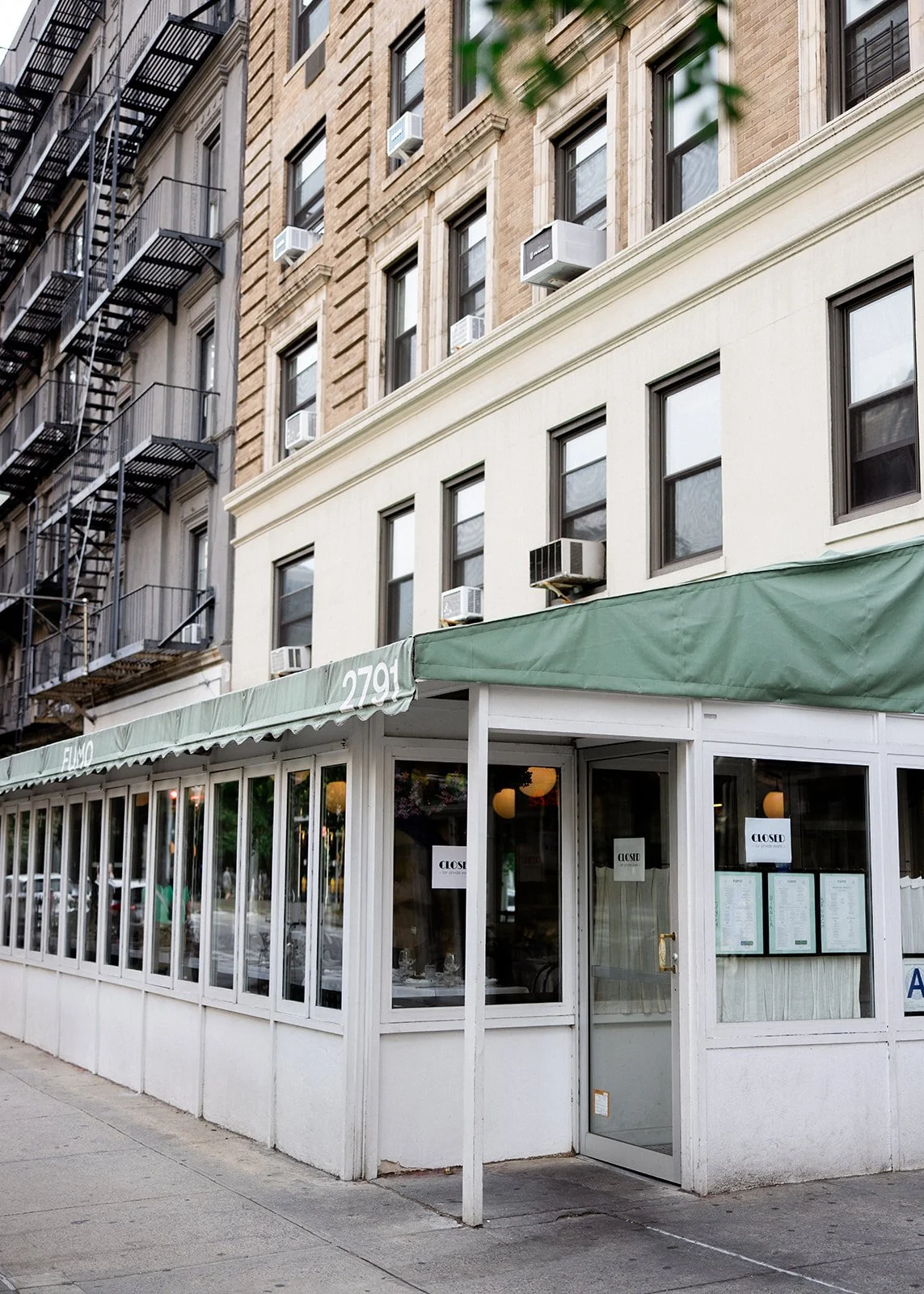Exterior view of a small restaurant or café with a green awning, large windows, and a glass door, located on the ground floor of a multi-story brick building in an urban area. Signs on windows indicate the restaurant is closed.
