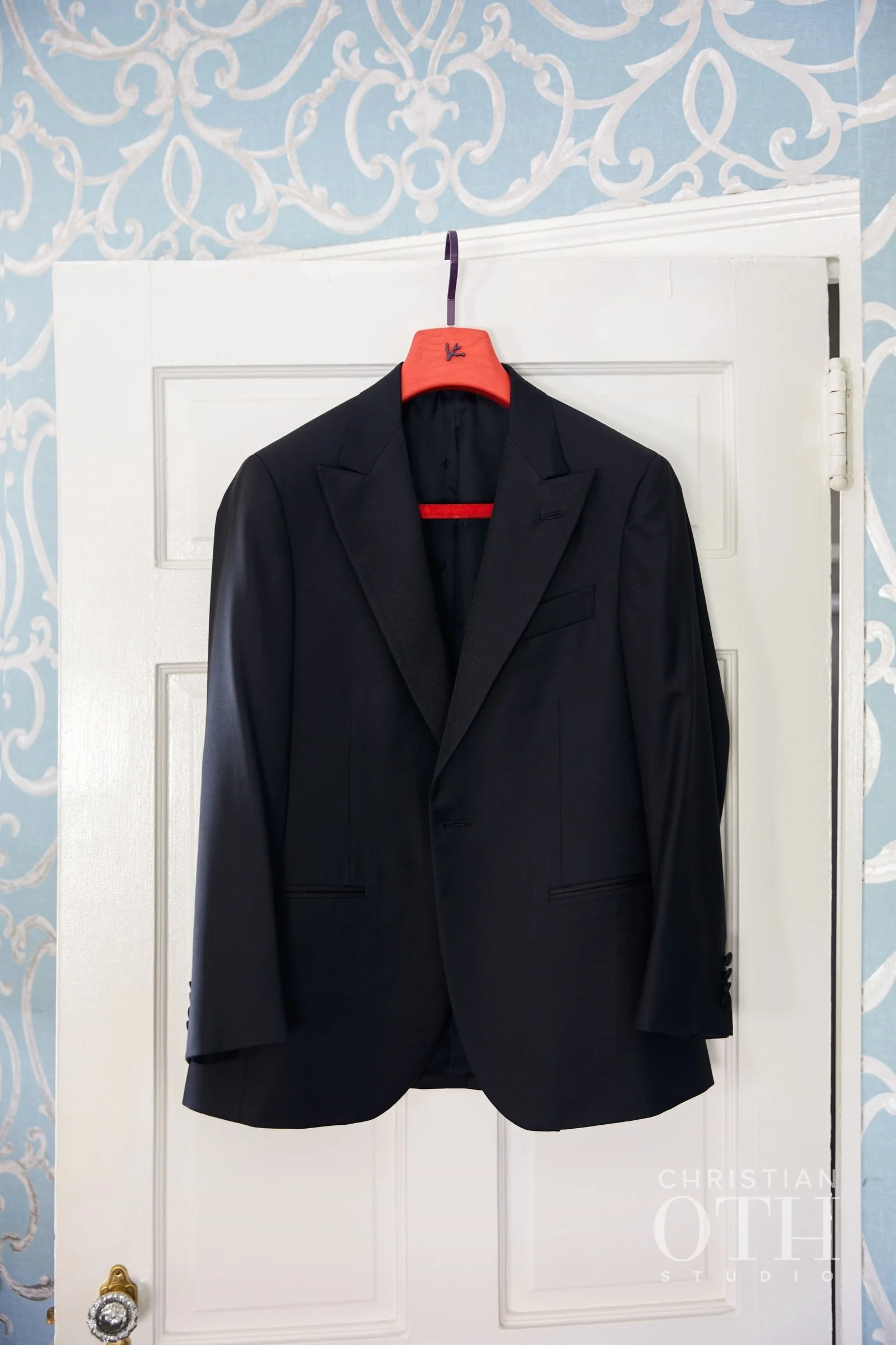 Black suit jacket hanging on a red hanger on a white door with blue wallpaper background.