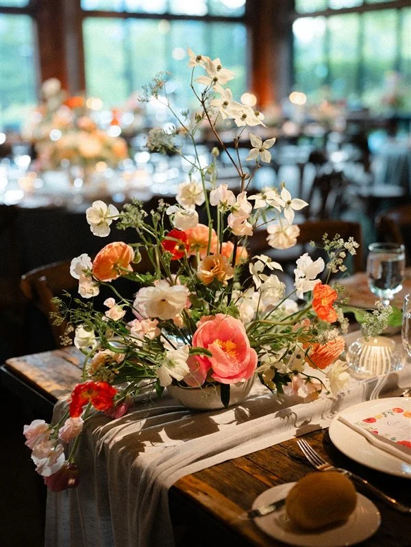 Elegant flower arrangement with pink, white, and peach blooms on a table setting at a formal event or restaurant, with blurred background of other tables and large windows