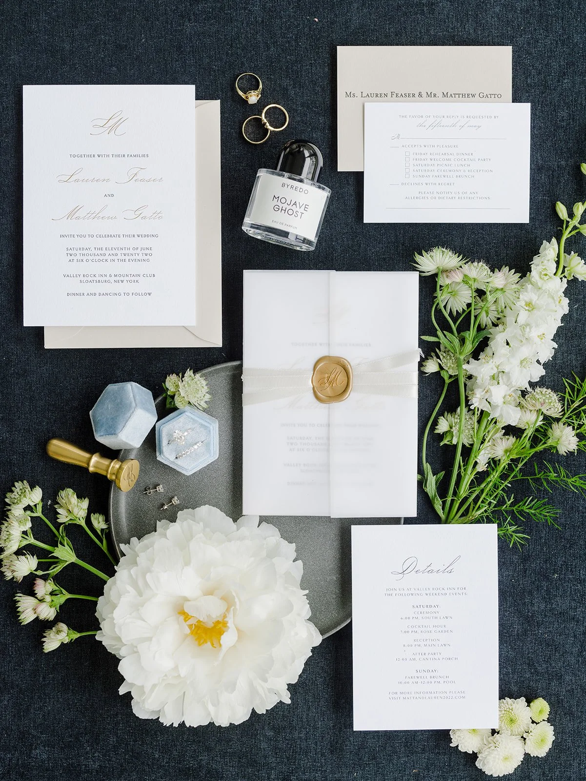 Wedding invitation and details suite with rings, perfume, and floral decor arranged on a dark surface.