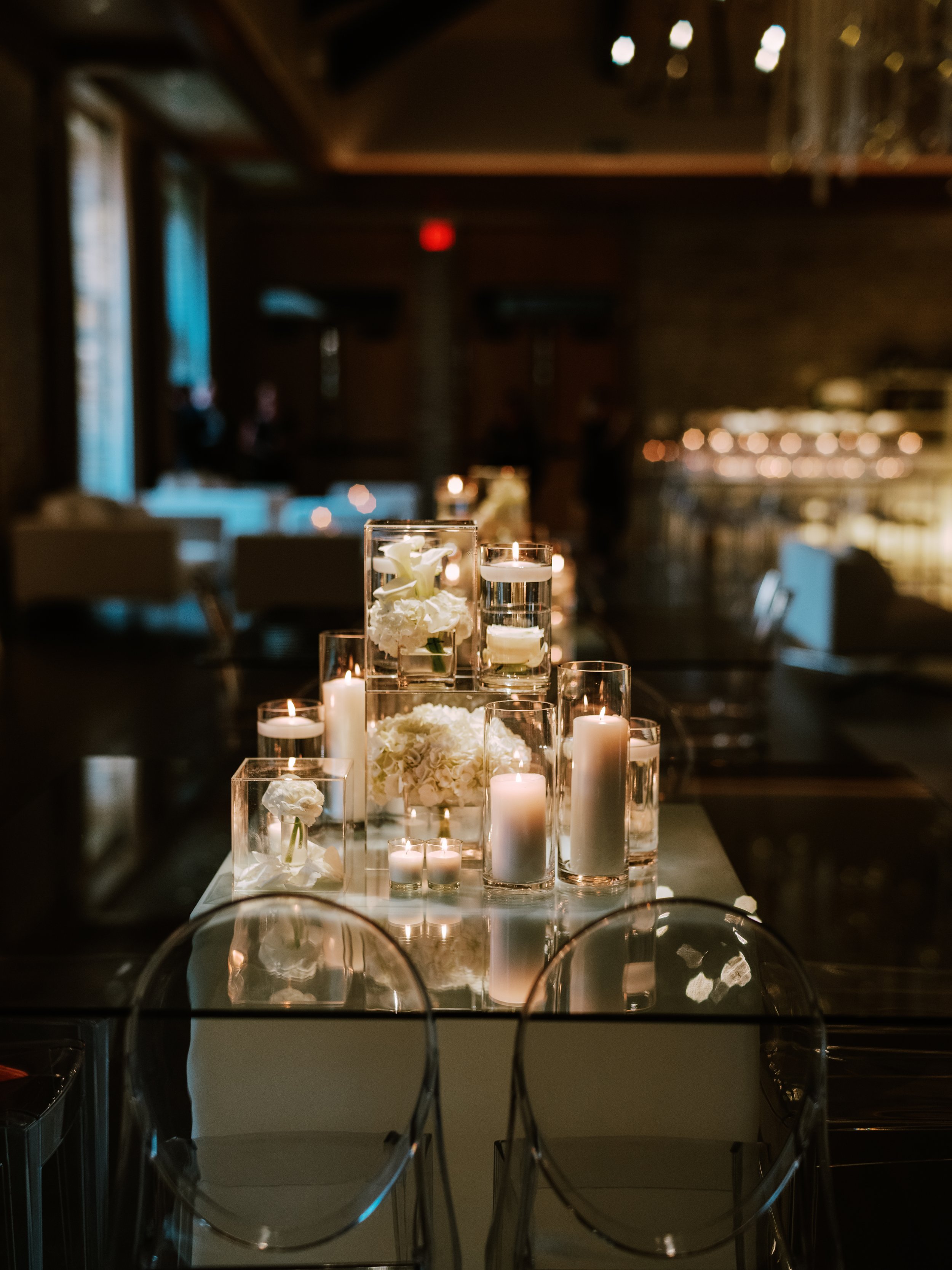 Table centerpiece with white flowers and candles in glass vases, set in a dimly lit room suitable for a formal event.