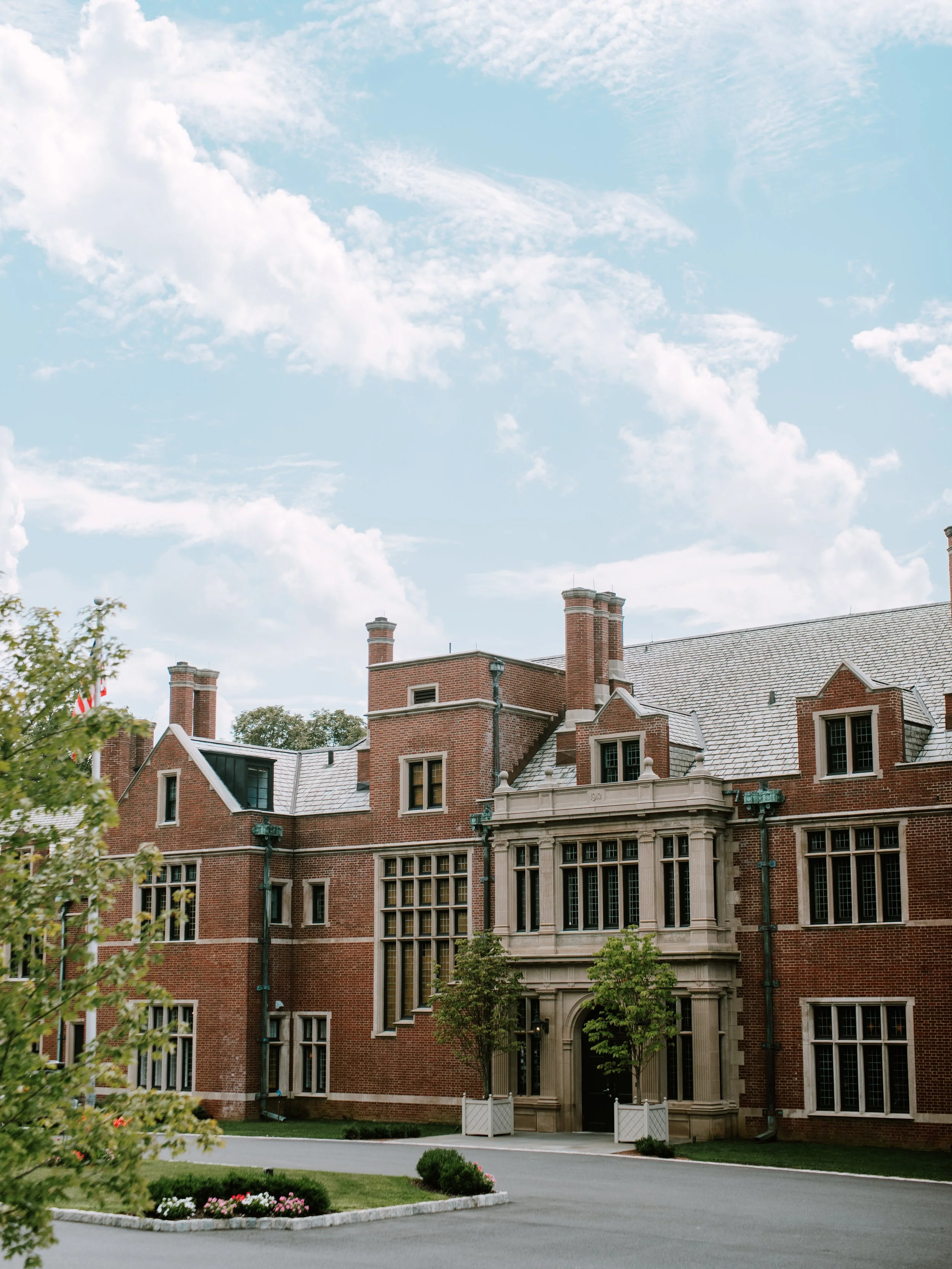 A large red brick mansion with multiple chimneys, tall windows, and a grand entrance with columns, surrounded by a green lawn, trees, and a street.