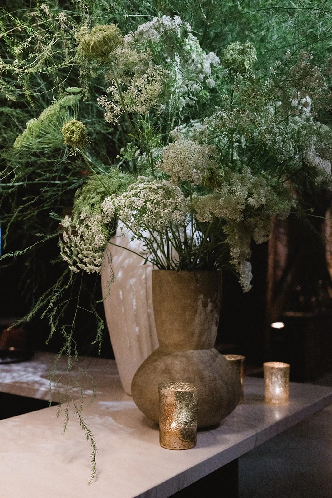 A large ceramic vase with white flowers and greenery, surrounded by three small gold-colored candle holders, on a white surface.