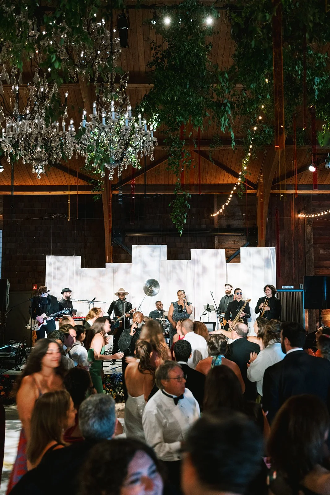 Band performing live on stage at an indoor event with chandelier, branches, and fairy lights, while guests dance and socialize.