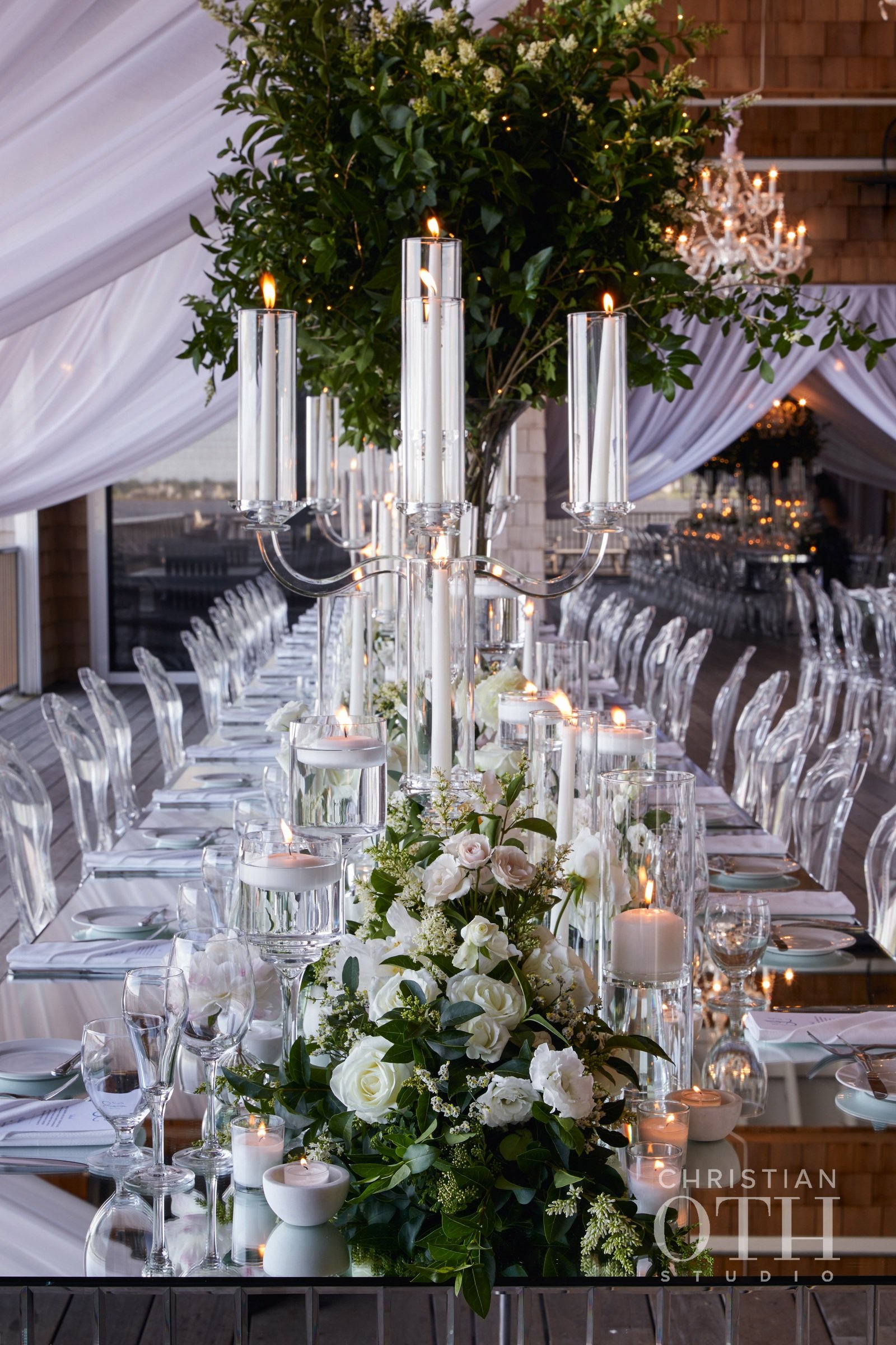 Elegant wedding reception table decorated with white roses, candles, and tall glass candelabras in a glass-topped venue with draped white fabric and a large leafy green arrangement.