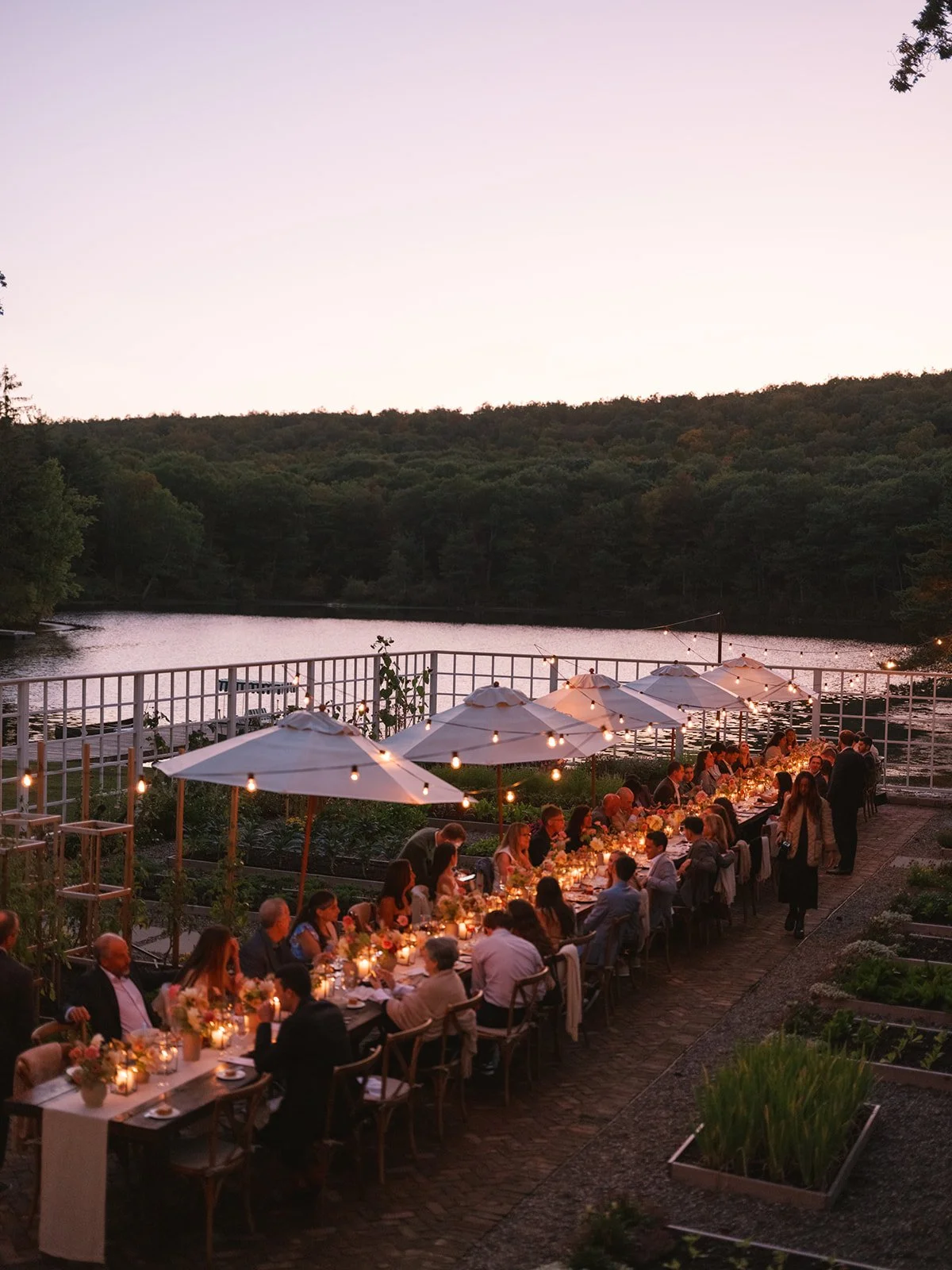 Outdoor dinner party by a river at sunset with string lights, umbrellas, and guests seated at long tables.