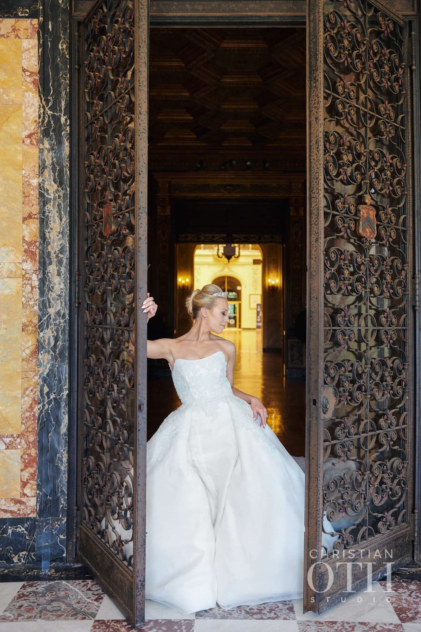 A bride in a white wedding gown standing partially behind ornate iron doors, looking down, with a regal interior background.