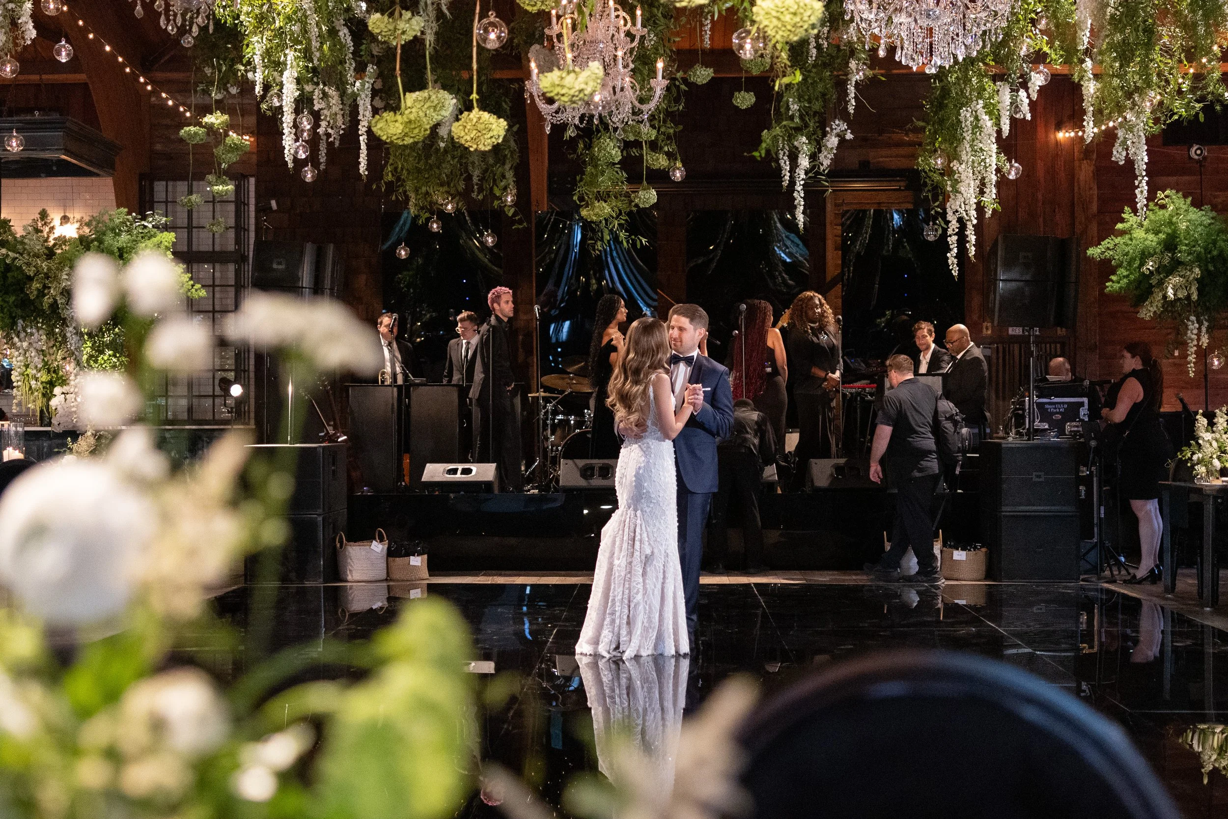 A bride and groom dancing in the center of a wedding reception hall, with a band playing and wedding guests in the background. The hall is decorated with hanging greenery, flowers, and crystal chandeliers.