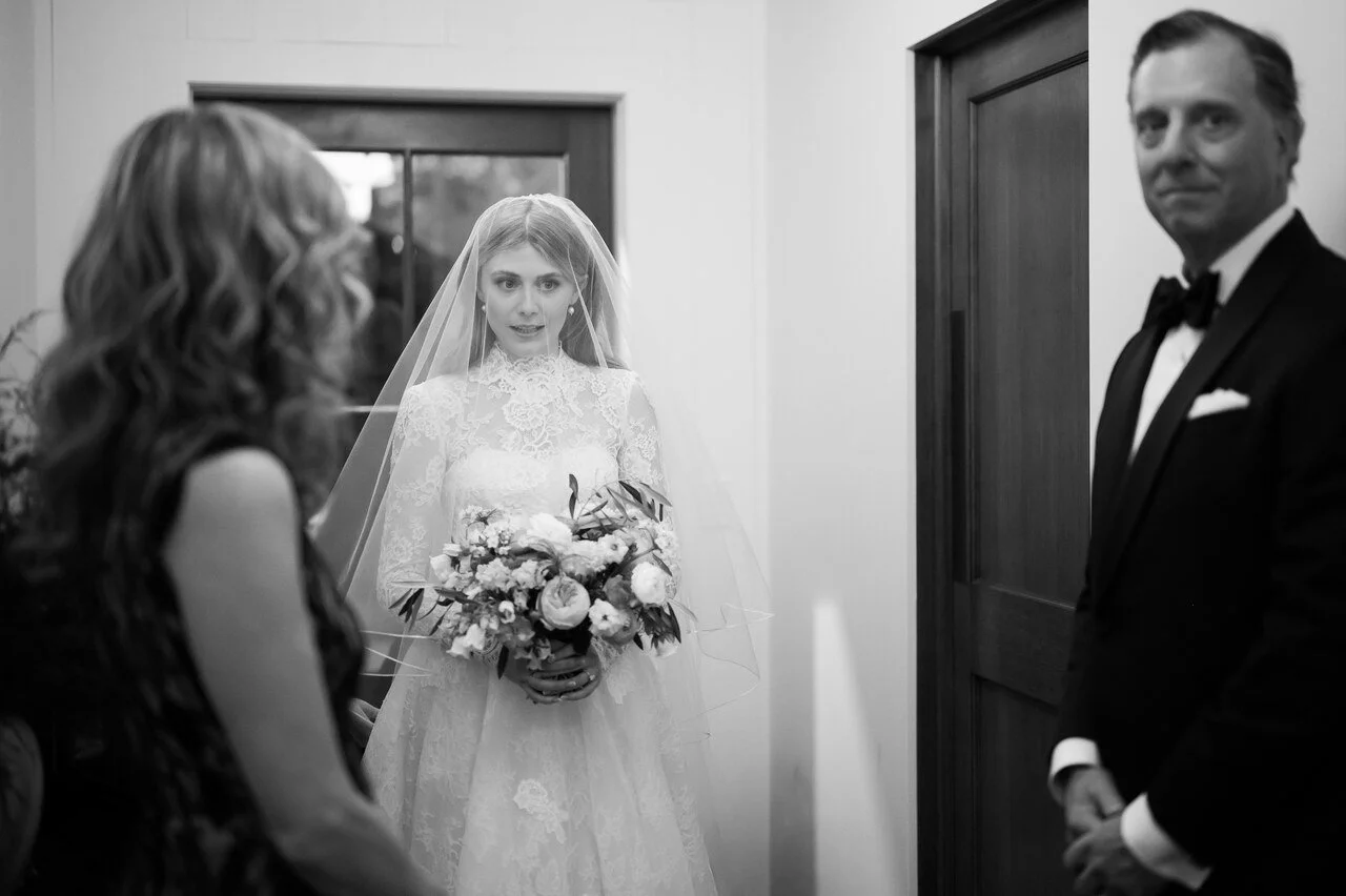 A bride in a lace wedding dress holding a bouquet, standing in front of a doorway, with a woman in a dark dress and a man in a tuxedo present.