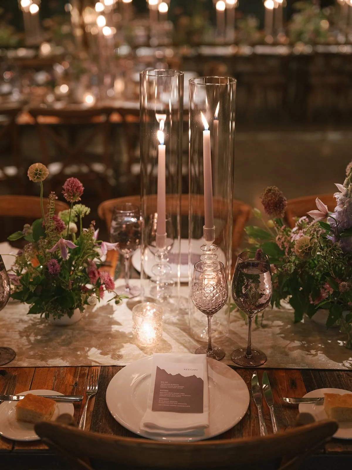 Elegant dinner table with floral centerpieces, tall glass candle holders with lit candles, and wine glasses, set in a warmly lit outdoor setting.