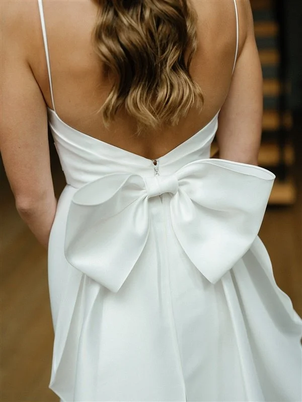 Back view of woman in white dress with large bow at waist and spaghetti straps, showing her long, wavy hair.