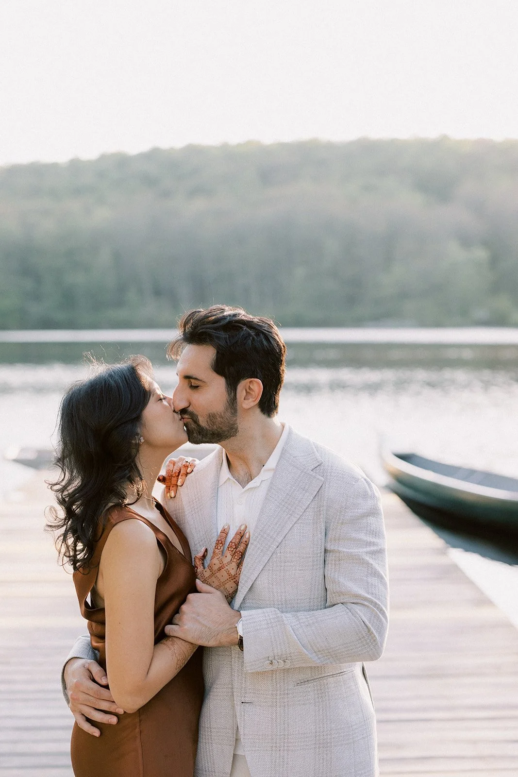 A couple kissing by a lake, with boats and a hill in the background.