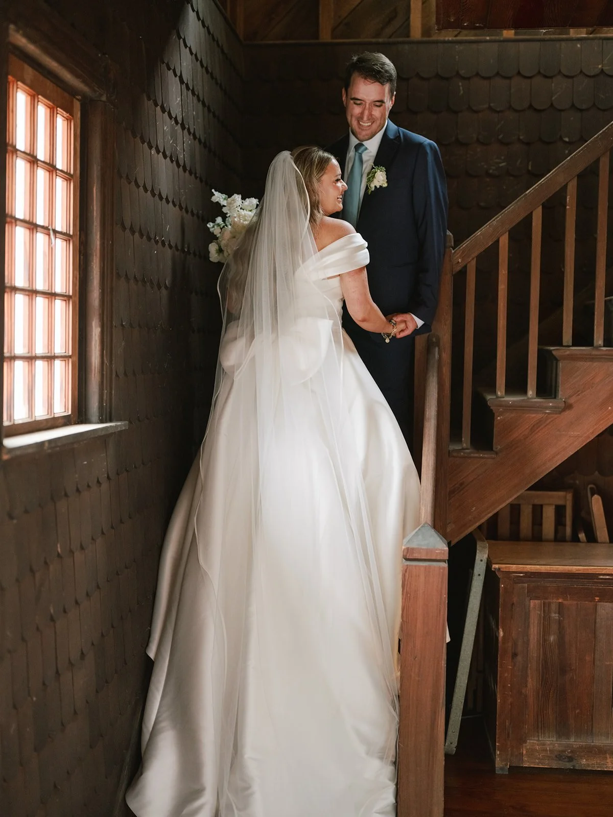 A bride and groom smile at each other on a staircase inside a wooden building during wedding. The bride wears a white wedding gown and veil, and the groom wears a dark suit with a light blue tie.