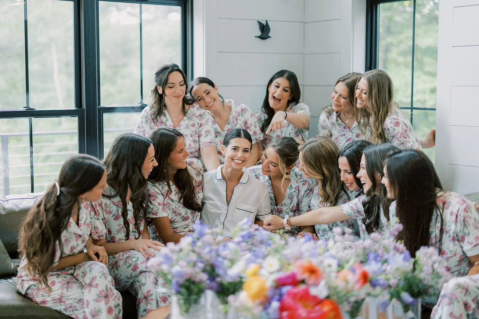 A group of women, wearing matching floral pajamas, are gathered around a woman in white pajamas, smiling and laughing together in a bright room with large windows and a floral arrangement in the foreground.