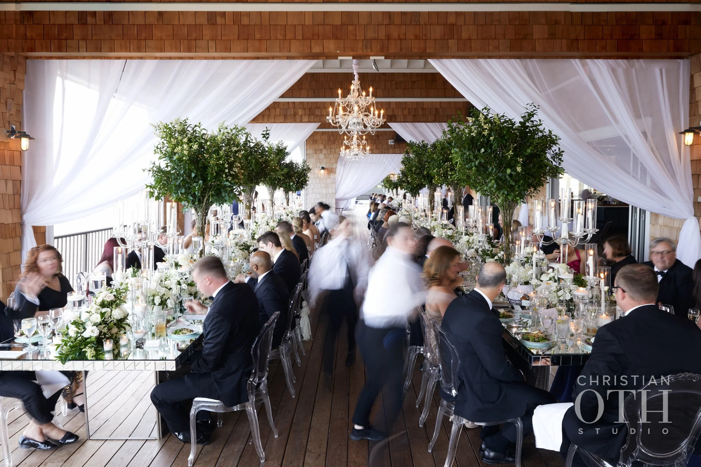 Elegant wedding reception with long banquet table, white floral centerpieces, tall candles, and trees for decoration in a wood-paneled venue, with curtains and chandeliers.