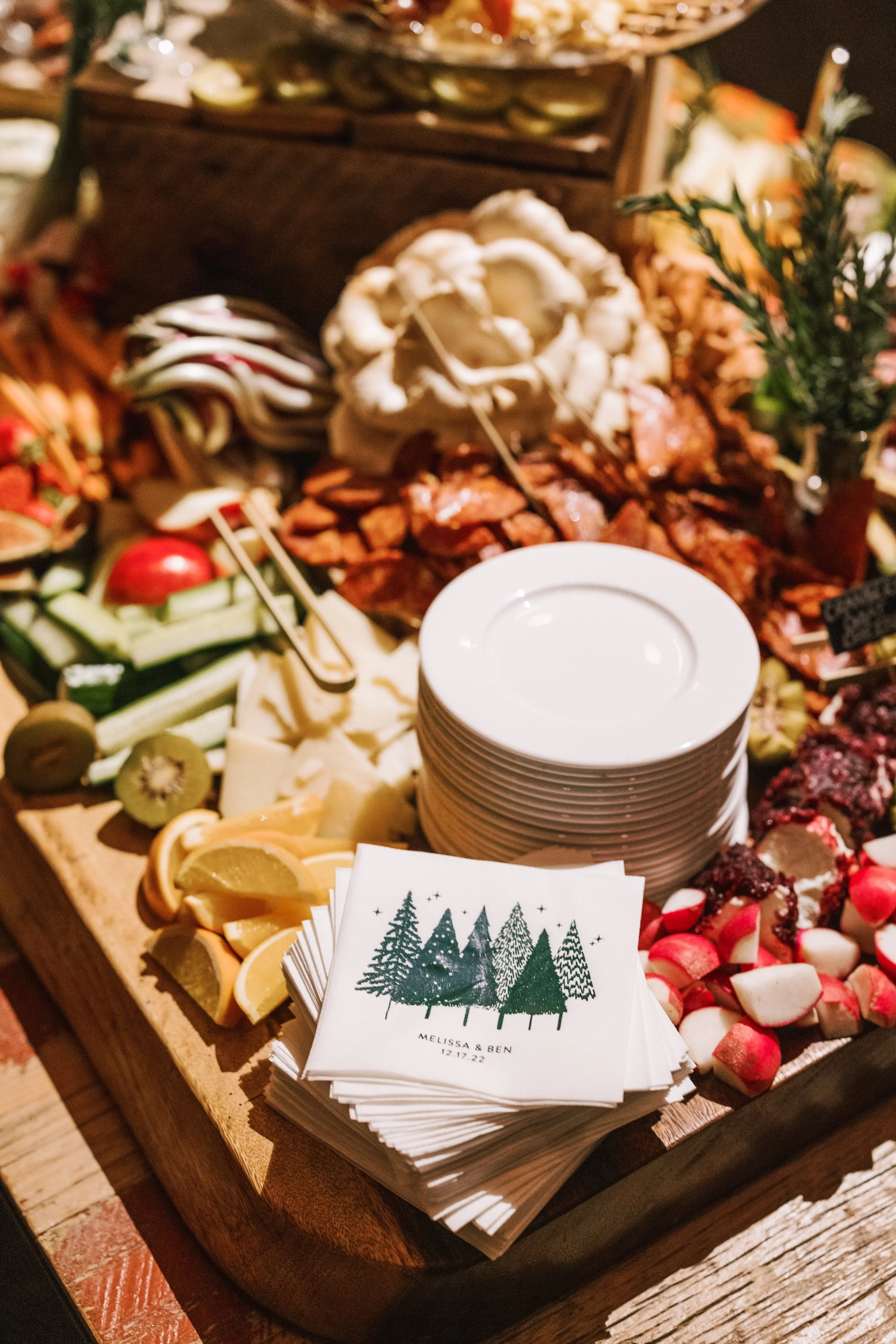 A wooden tray with a variety of sliced fruits, vegetables, and cheeses, alongside white napkins with a Christmas tree design and the names Melissa and Ben, date 12.17.22, and a stack of white plates.