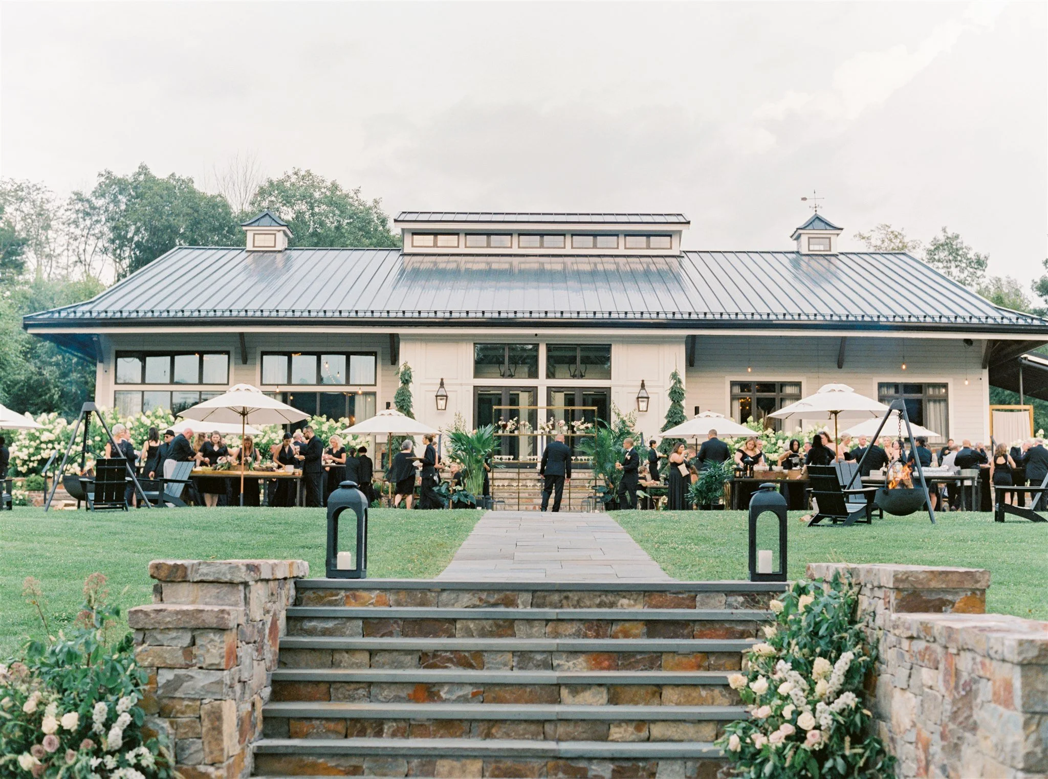 A wedding reception outdoor event with guests, tables, umbrellas, and a large house with a dark roof in the background.