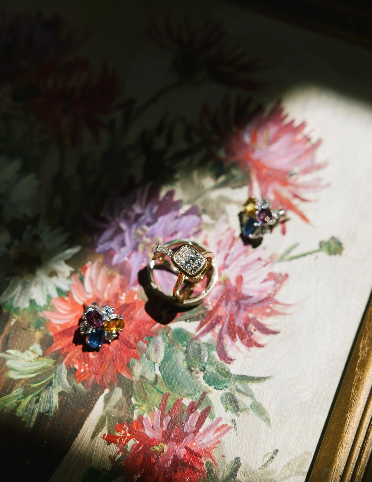 Close-up of jewelry, including a diamond ring and colorful gemstone earrings, placed on a floral painting with pink, purple, and white flowers.