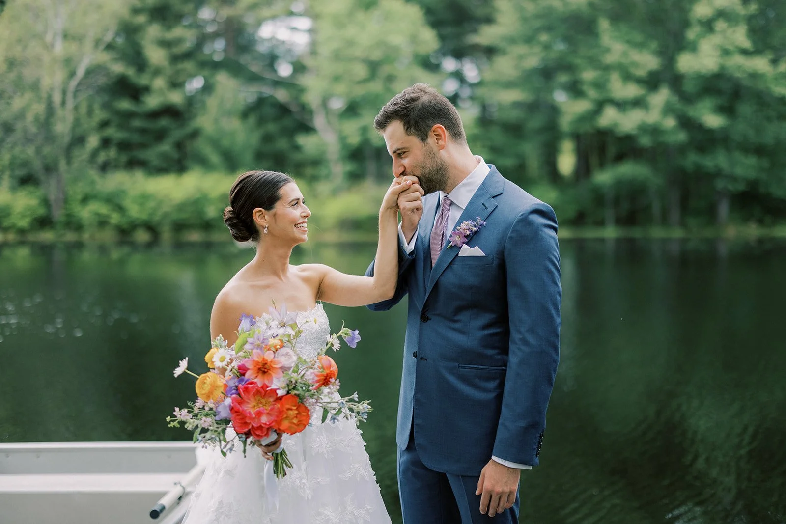 A bride in a white wedding dress holding a colorful bouquet of flowers, and a groom in a blue suit standing by a lake, sharing a tender moment with the bride touching the groom's face.