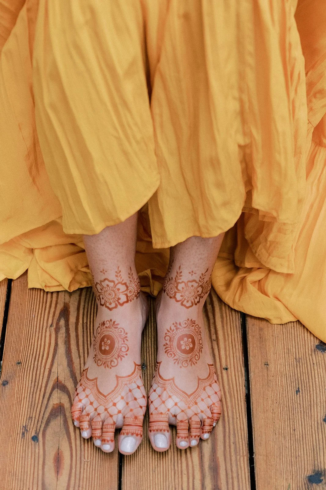 Feet decorated with henna, wearing a yellow dress, on a wooden floor.