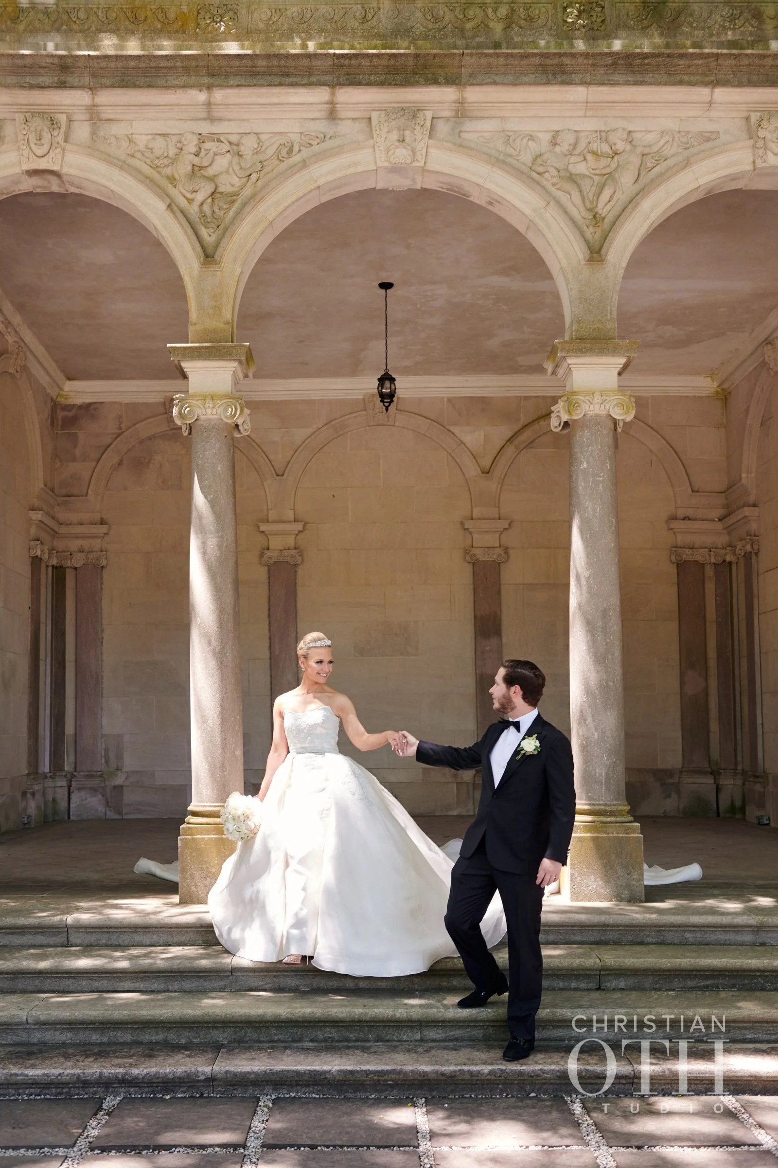 A bride and groom holding hands on the steps of a historic building with classical columns and ornate carvings, during their wedding photoshoot.
