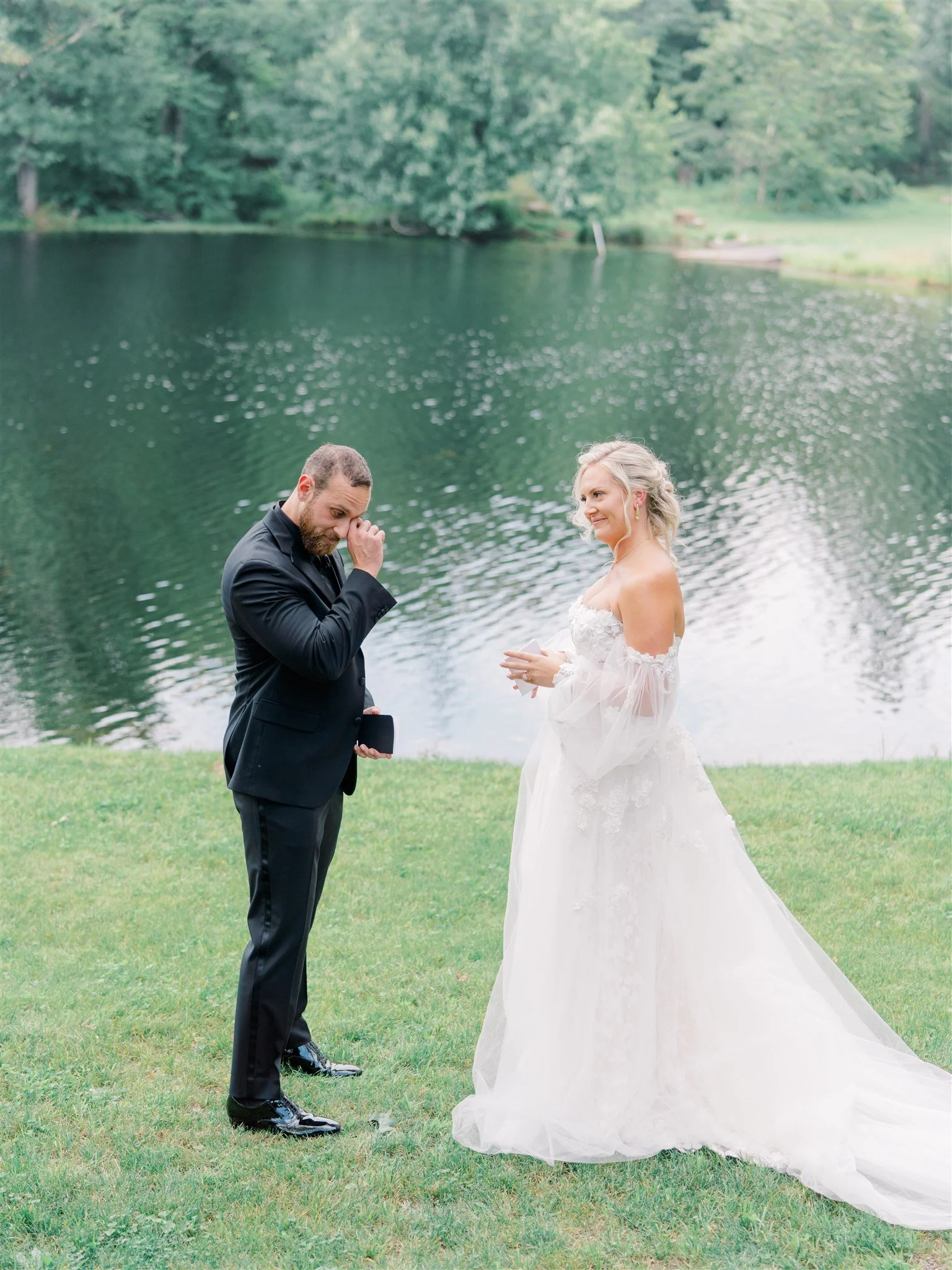 A couple getting married outdoors by a lake, the groom appears emotional while the bride smiles