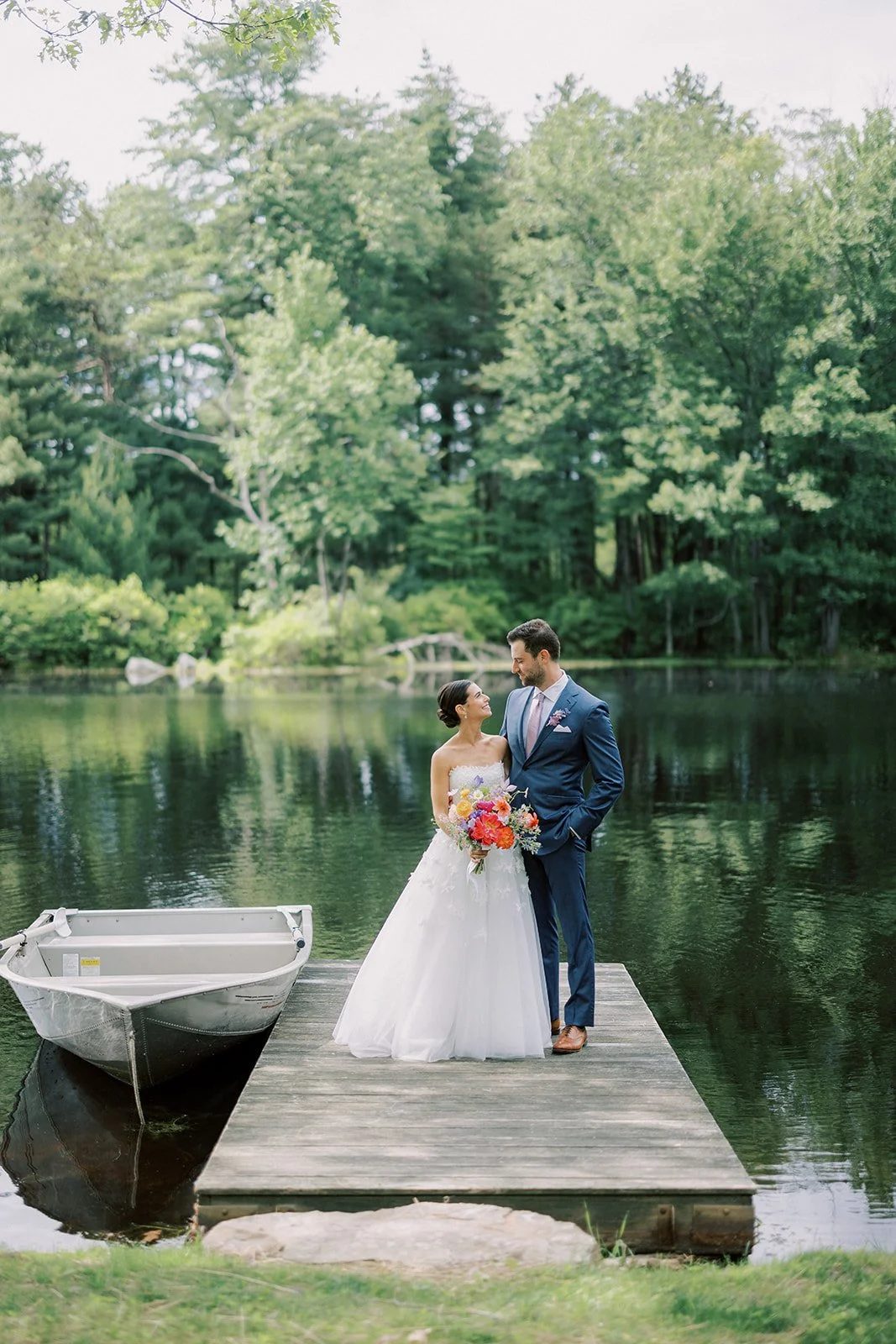 A bride and groom stand on a wooden dock by a lake, with a boat tied to the side. They are gazing into each other's eyes surrounded by lush green trees.