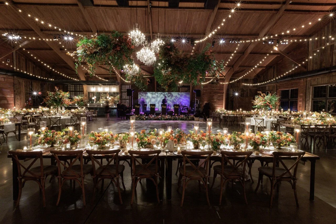 A decorated wedding reception venue inside a rustic wooden barn, featuring long tables with flower arrangements and candles, string lights, and a stage with music equipment.