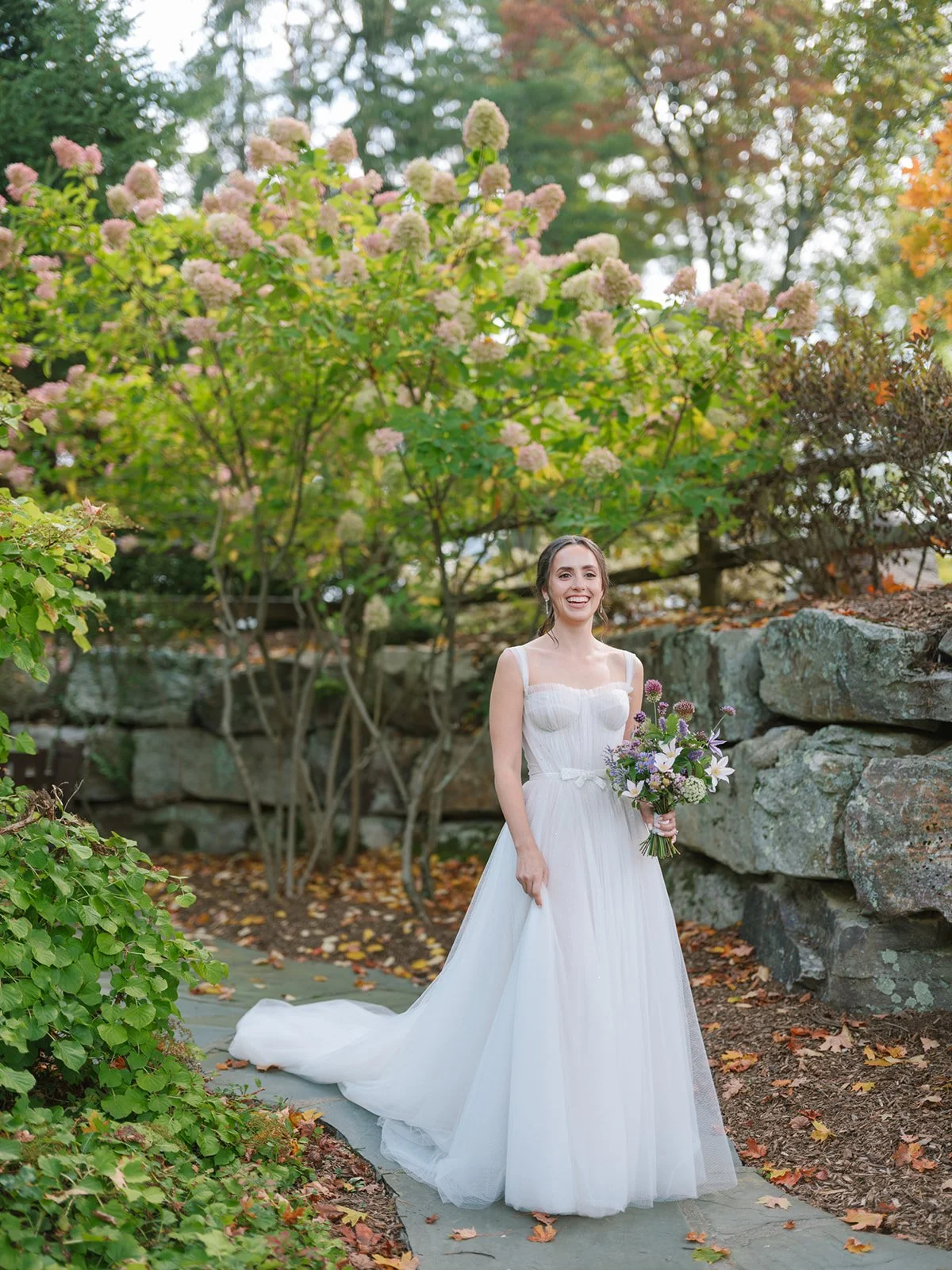 A smiling bride in a white wedding dress holding a bouquet of flowers outdoors, surrounded by green and autumn-colored foliage.