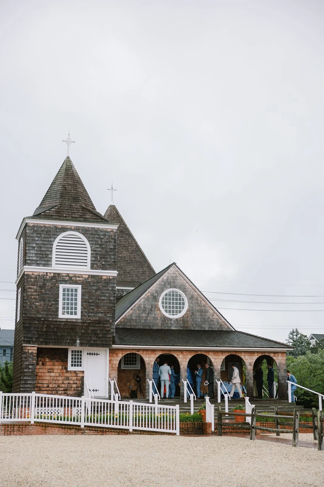 People gathered in front of a wooden church with a steep roof and crosses on top, overcast sky.