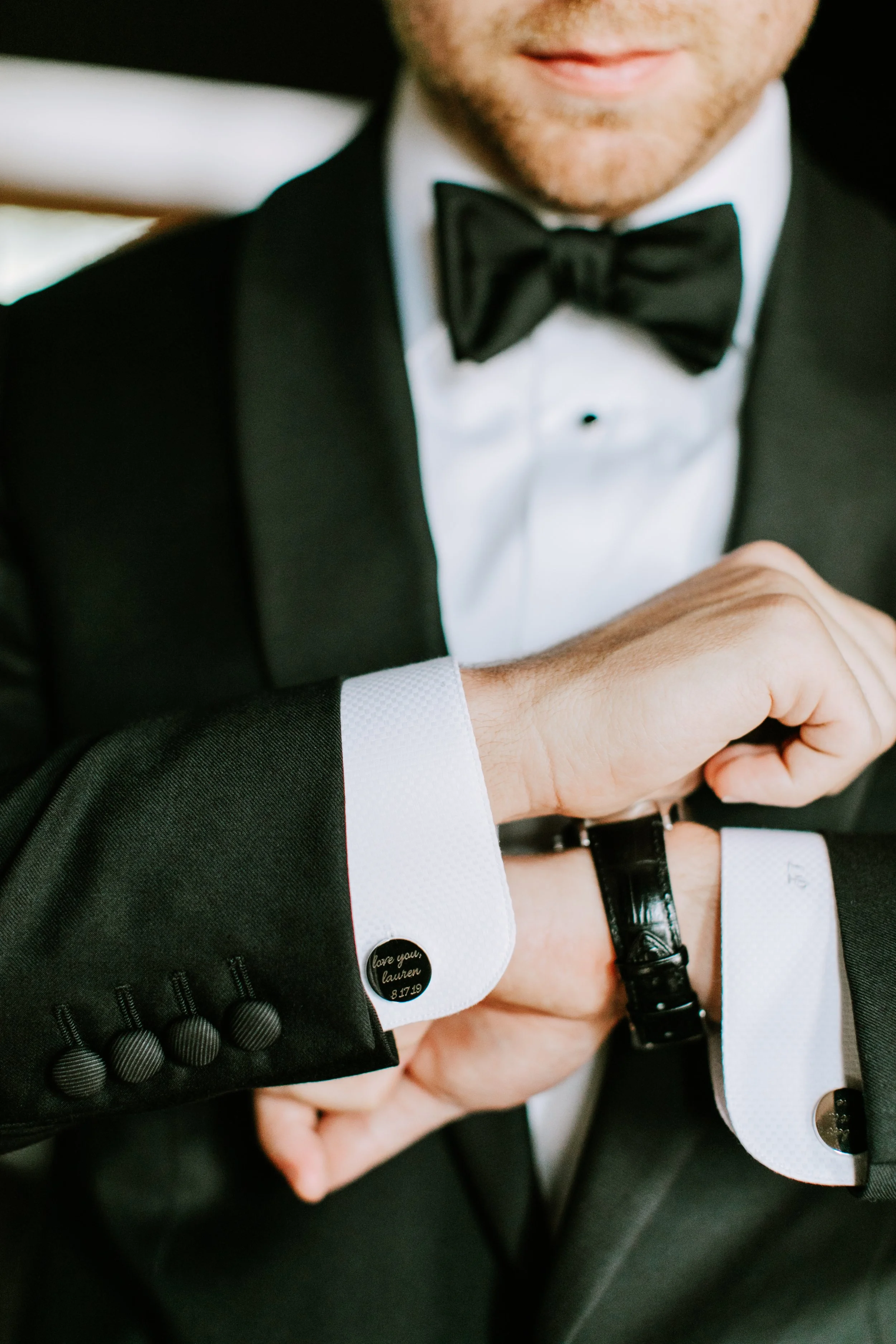 Close-up of a man in a tuxedo adjusting his watch, wearing a white shirt with French cuffs, cufflinks, and a black bow tie.