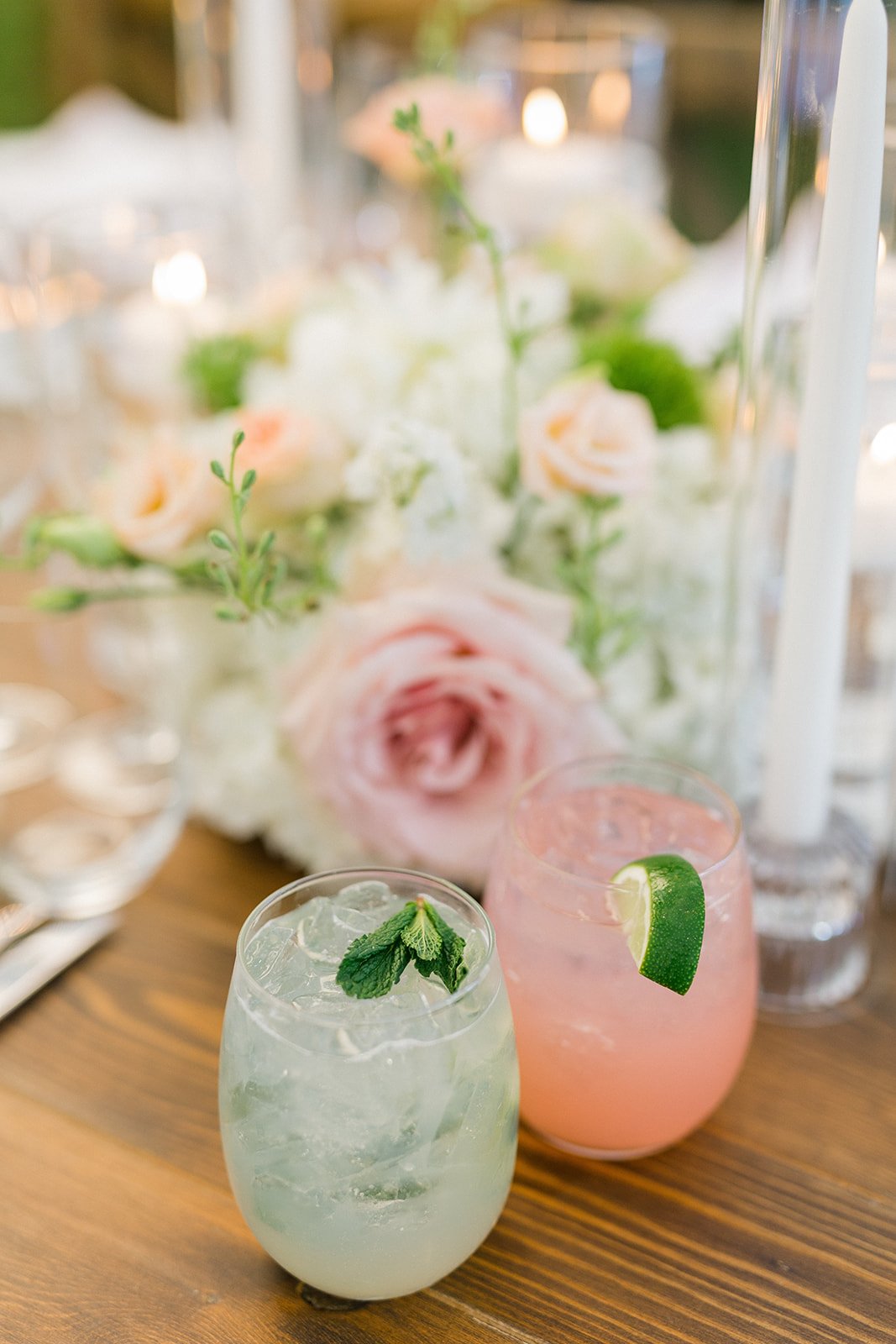 Two cocktails, one with mint and ice and one with lime, on a wooden table with a floral centerpiece in the background.