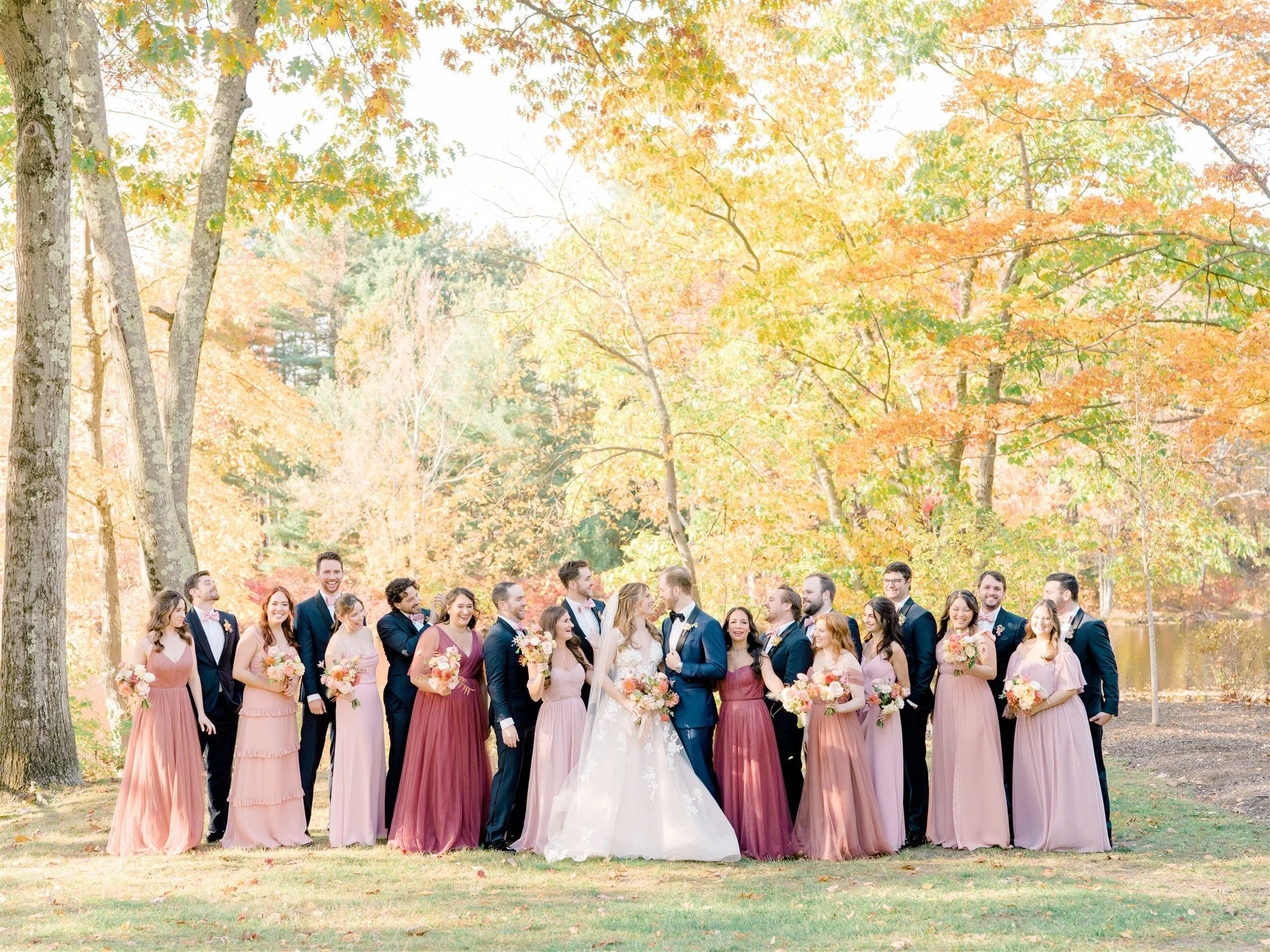 A wedding party with bridesmaids in pink and purple dresses and groomsmen in dark suits, standing outdoors under autumn-colored trees near a lake.