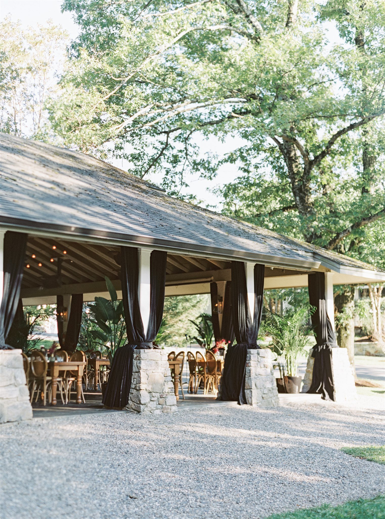 An outdoor pavilion with stone pillars and black curtains, surrounded by green trees and chairs inside, set for an event or gathering.