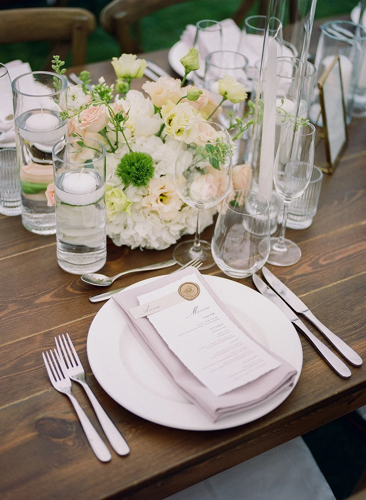 A formal table setting with a white plate, silverware, a folded napkin with a menu on top, and glassware including wine and water glasses. The centerpiece is a floral arrangement of white and blush flowers with greenery, with candles in tall glass ho