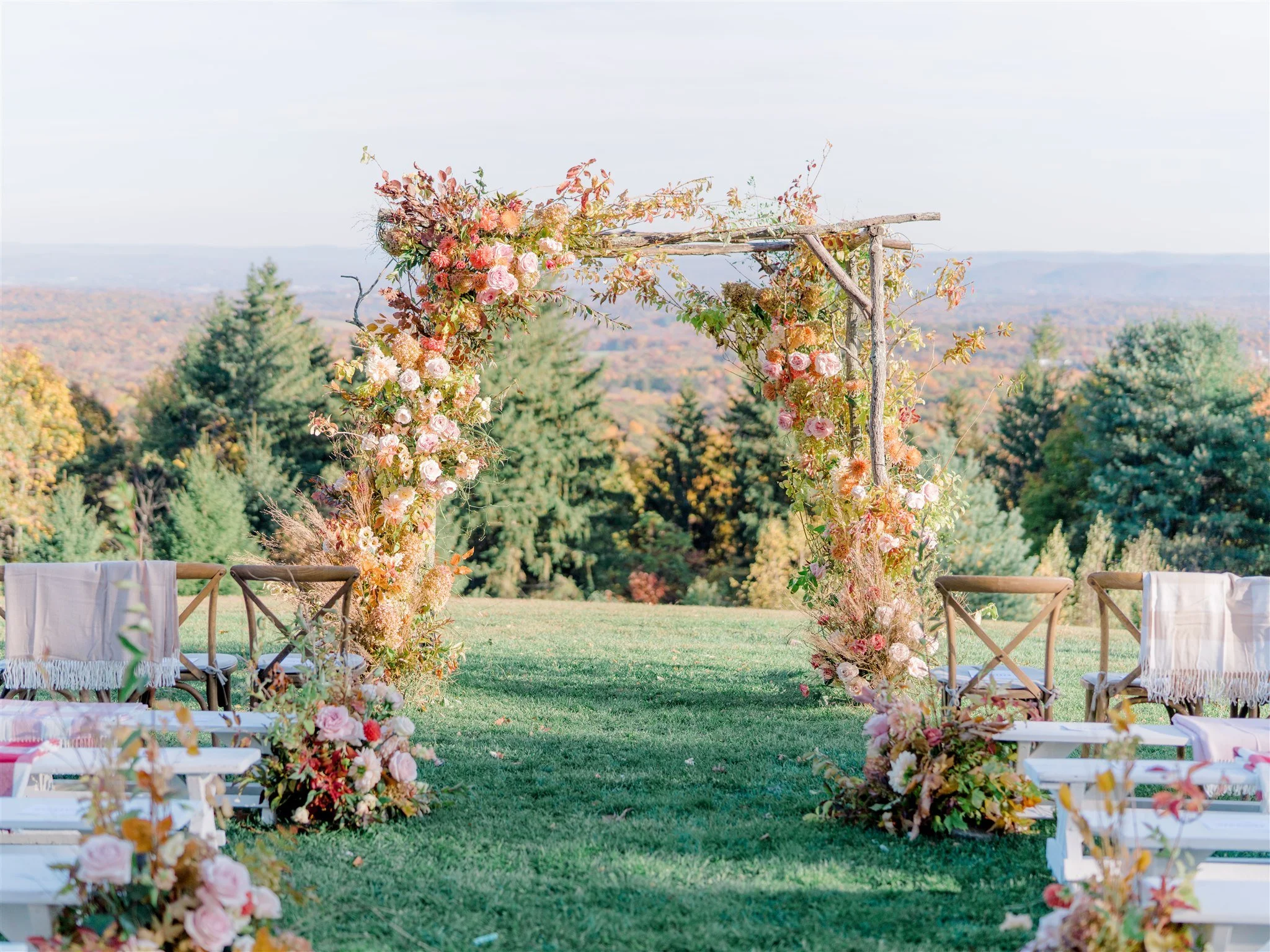 An outdoor wedding arch decorated with pink and white flowers, greenery, and branches, set on a grassy area with chairs nearby, overlooking a scenic landscape with trees and mountains.