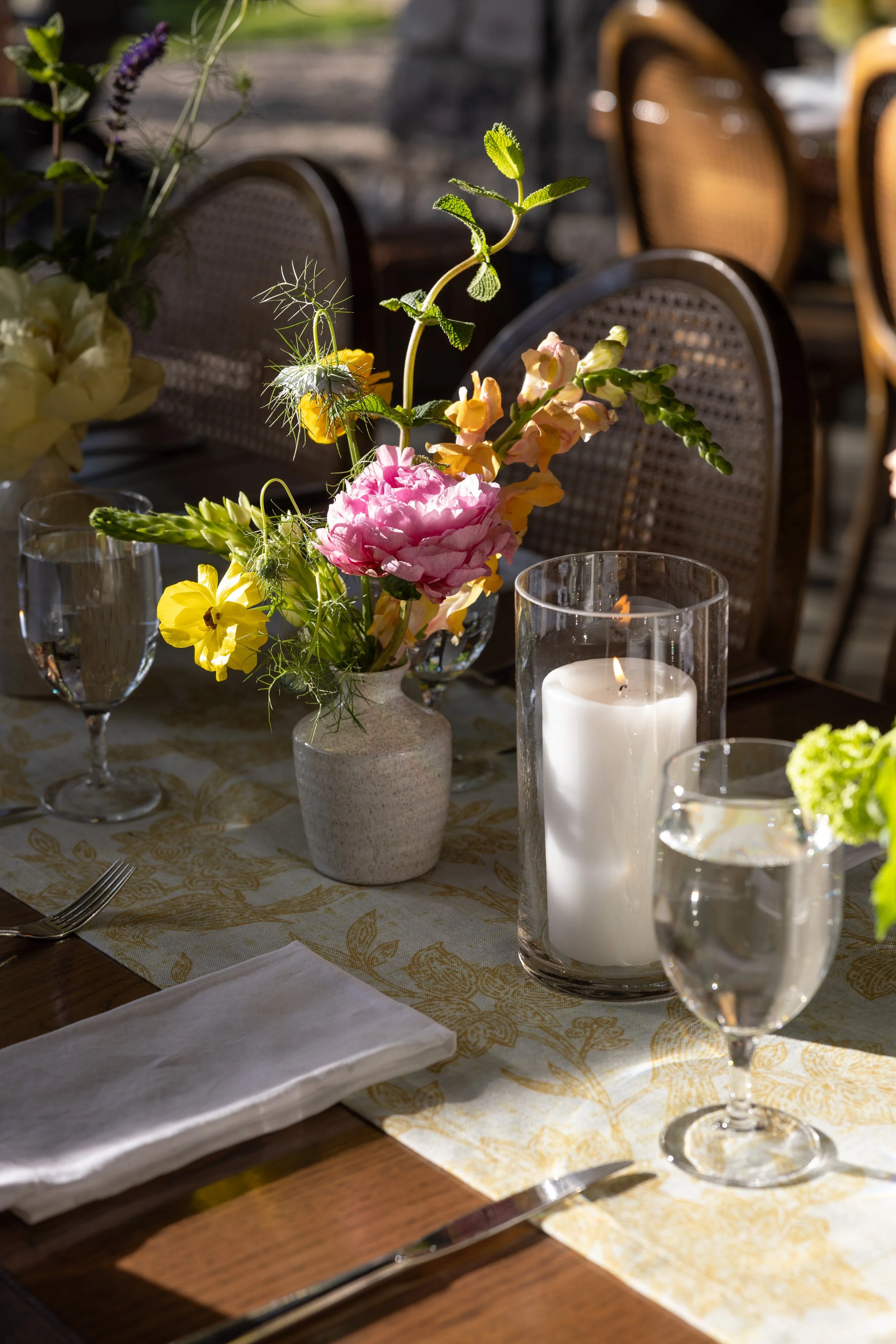 A dining table decorated with a pink flower arrangement, a lit white candle in a glass holder, two glasses of water, and a folded white napkin, set in a well-lit outdoor setting with chairs and other table settings visible in the background.
