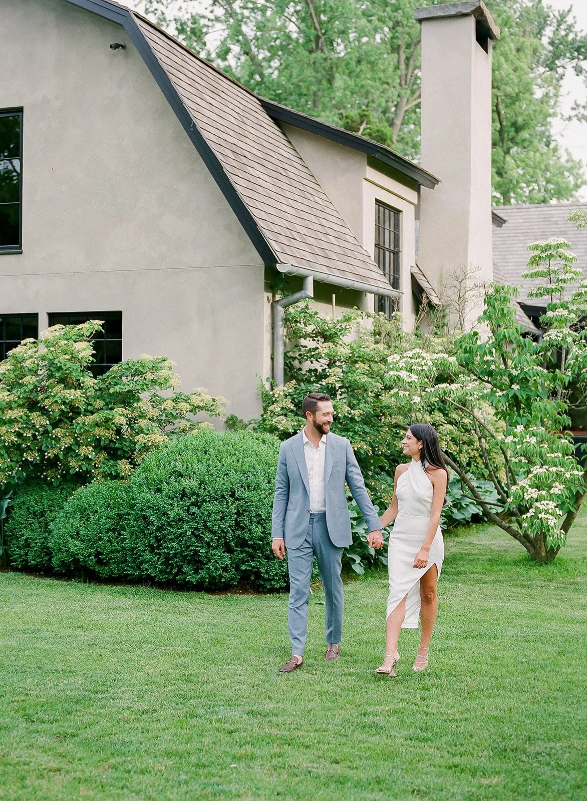 A smiling couple holding hands and walking across a green lawn in front of a beige house with lush bushes and trees in the background.