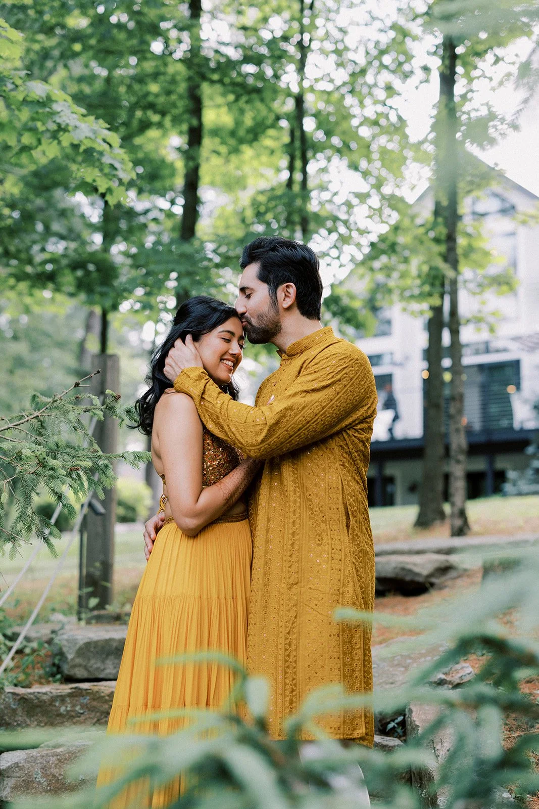 A couple dressed in traditional Indian clothing sharing a tender moment outdoors among trees, with the man gently holding the woman's head as they smile and close their eyes.