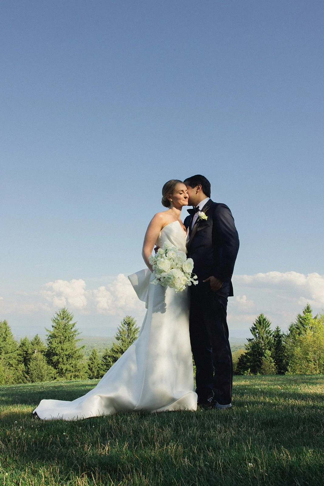 A bride and groom stand close together on a grassy field under a blue sky, sharing a tender moment. The bride is in a white wedding gown holding a bouquet of white flowers, and the groom is in a black tuxedo with a white boutonniere.